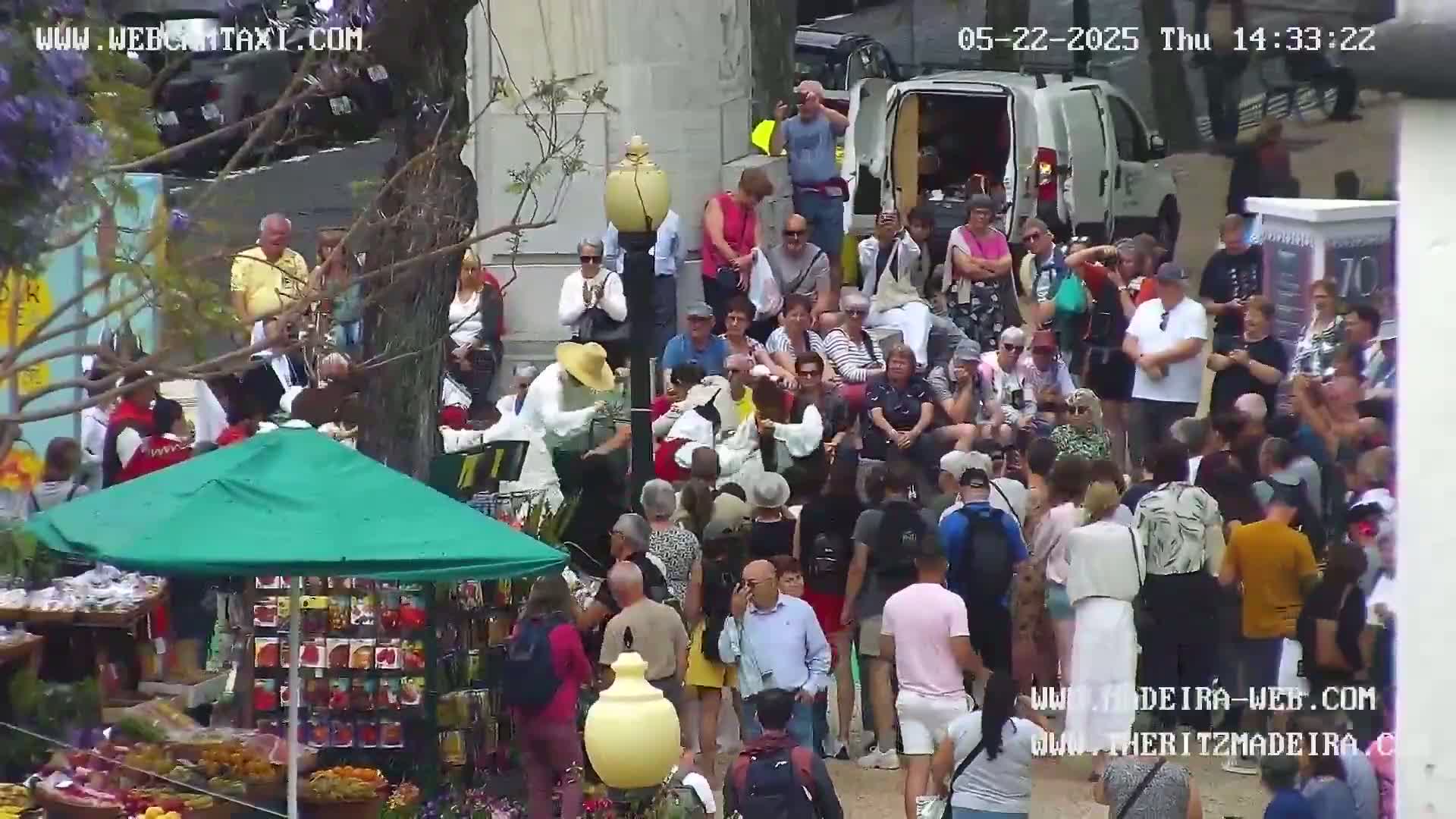 A large crowd of people watches two performers in white costumes on a sunny day in a town square, near a produce stand under a green umbrella.
