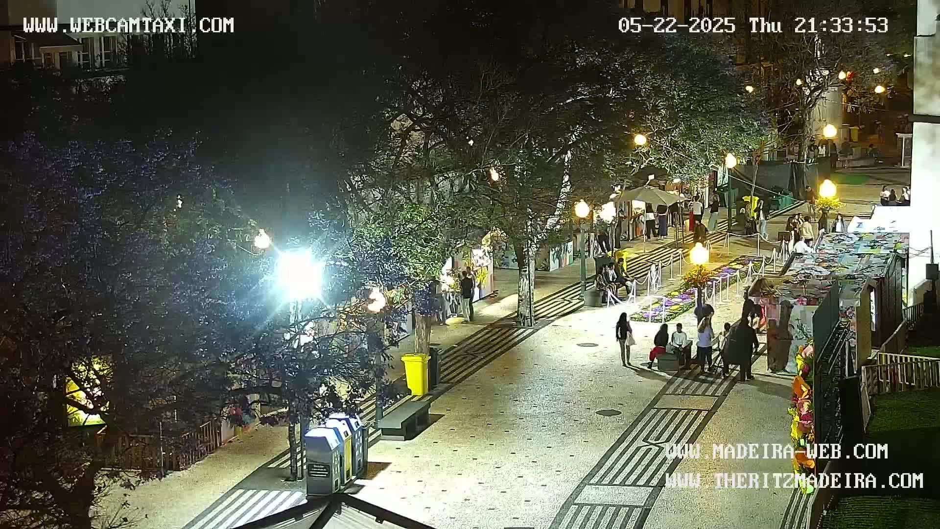 A nighttime, outdoor scene shows a paved pedestrian area lined with trees and small stands, with people strolling along under bright lights.