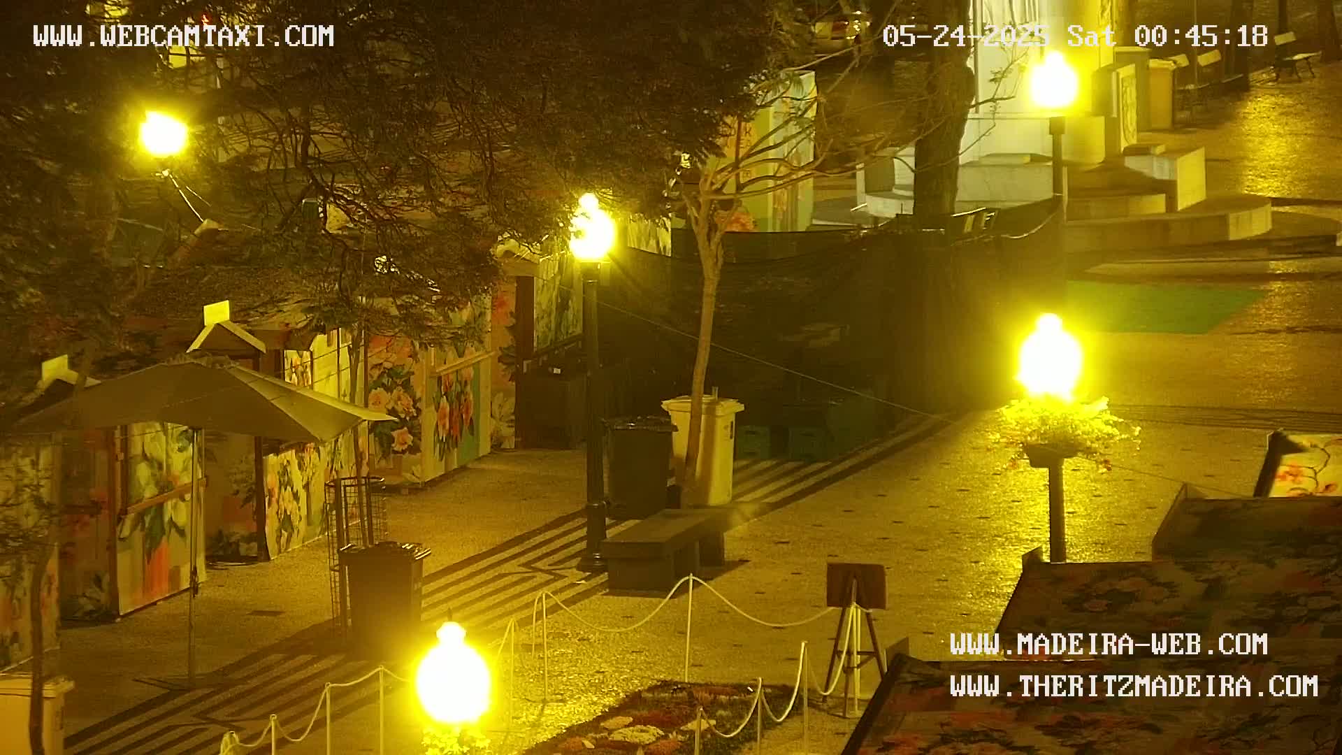 A nighttime view of an outdoor plaza with several illuminated structures, small stalls with painted sides, and string lights, all set on a patterned pavement.