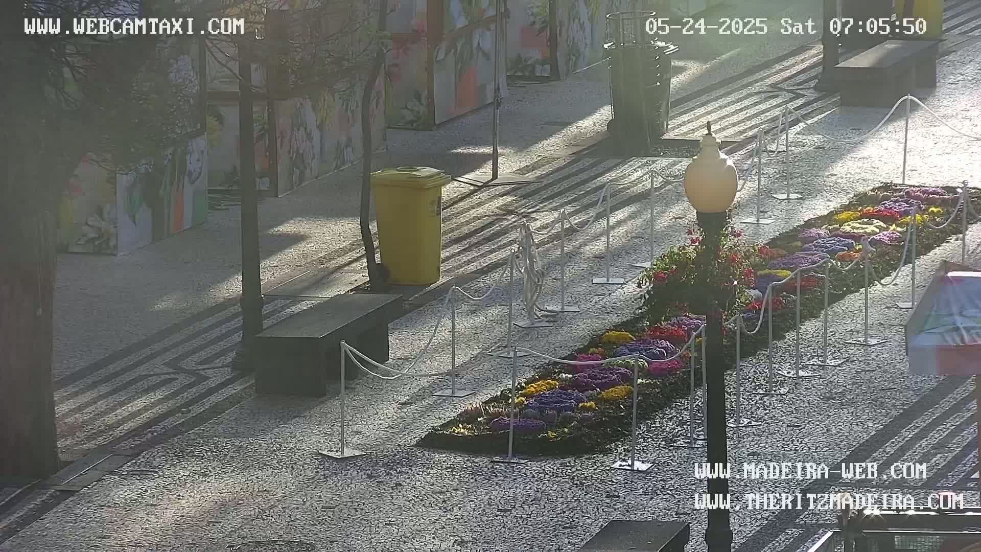 A sunny outdoor paved area features a long, colorful flower bed bordered by white stanchions, a yellow trash can, and a lamppost.
