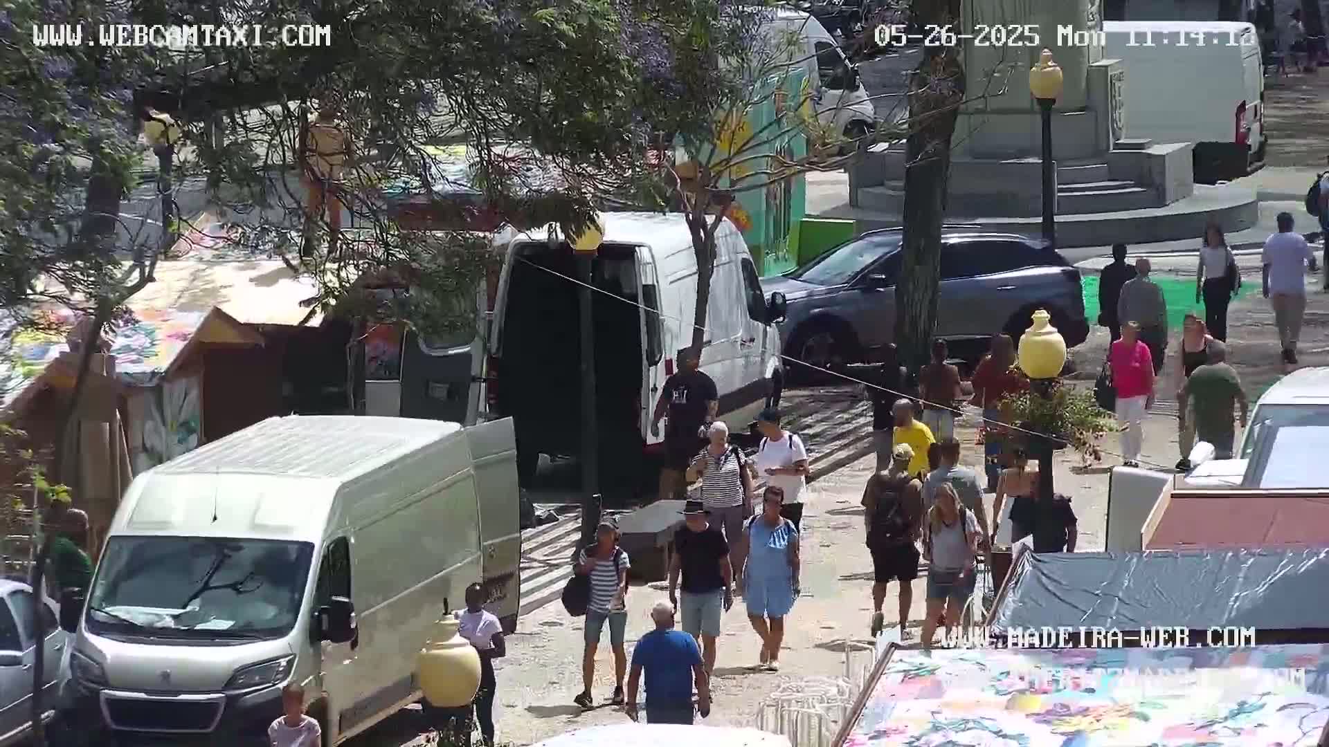 A sunny outdoor scene shows people walking among parked vans and market stalls.