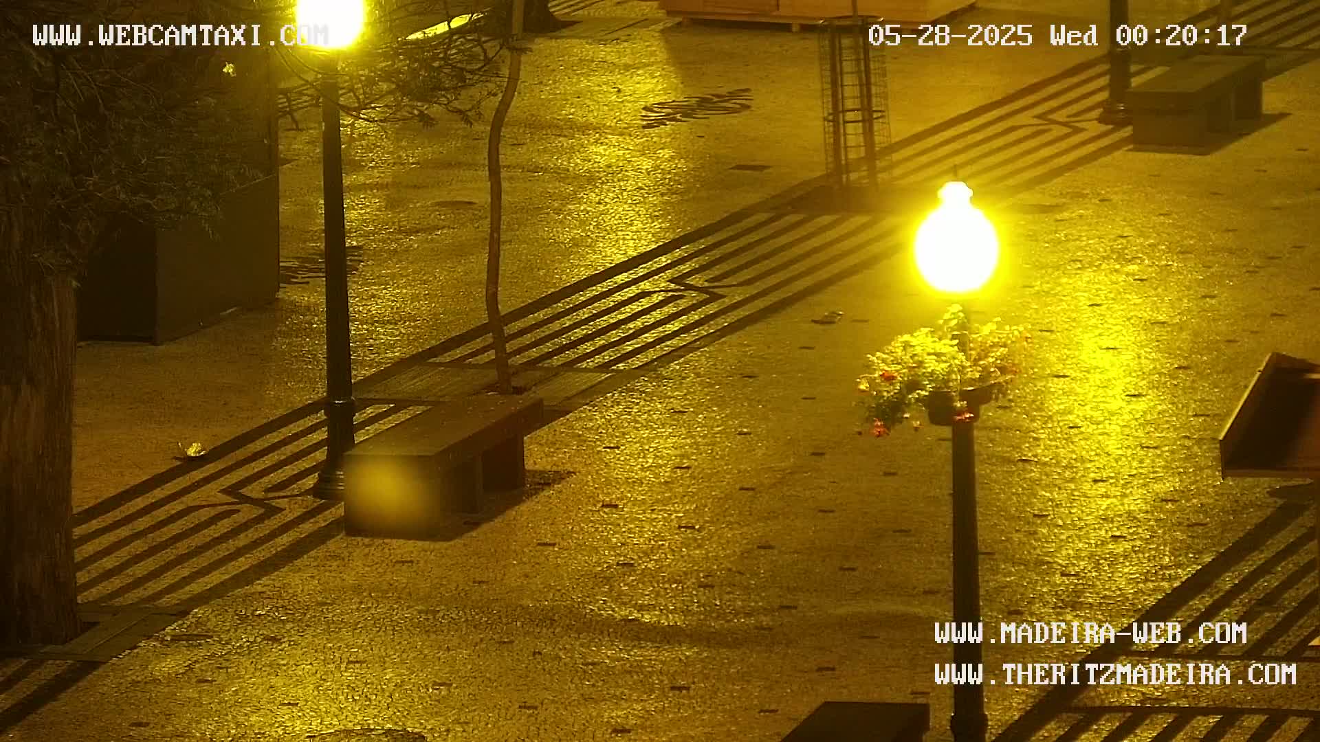 A wet, paved plaza is illuminated by a streetlamp at night, casting shadows from benches and railings onto the glistening surface.