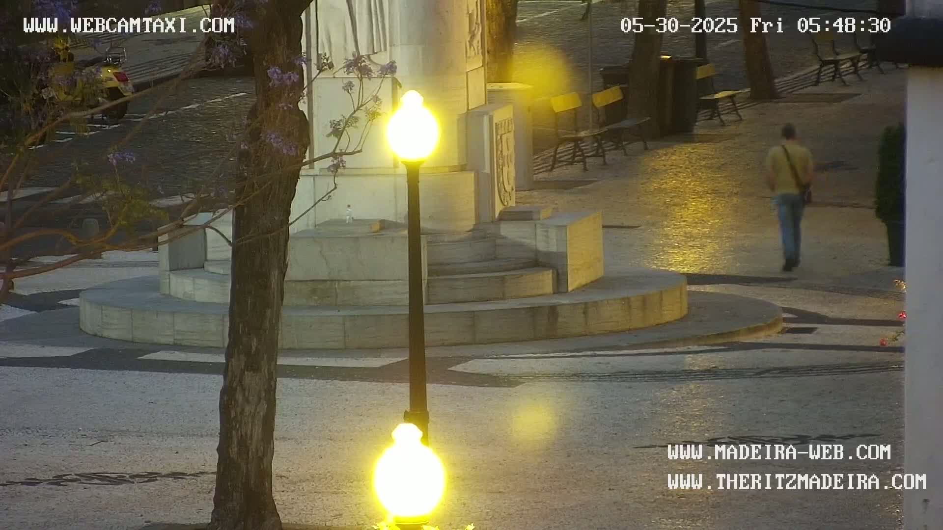 A man walks across a plaza at dusk, past a stone monument and a lit lamppost, under a mostly clear sky.