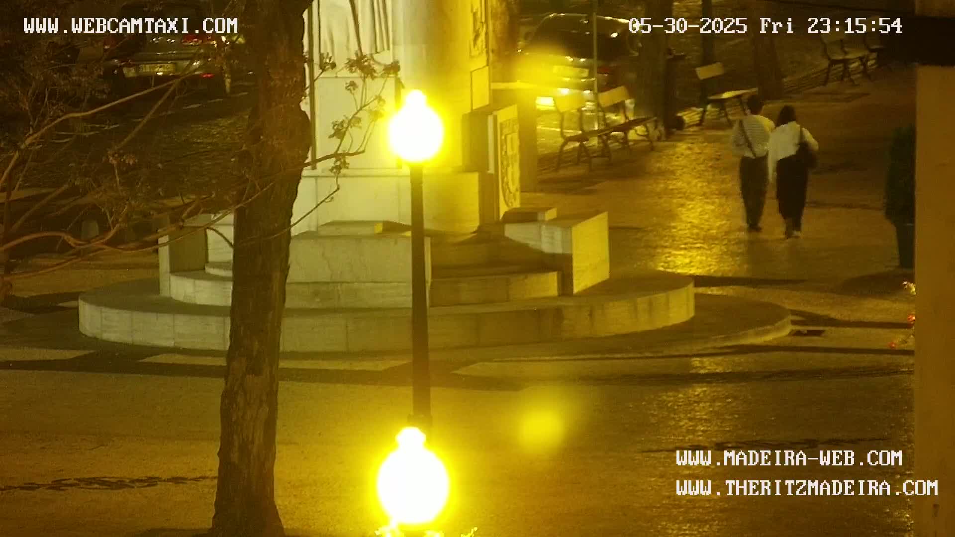 A nighttime scene shows two people walking past a stone monument and street lamps in a wet, paved area.