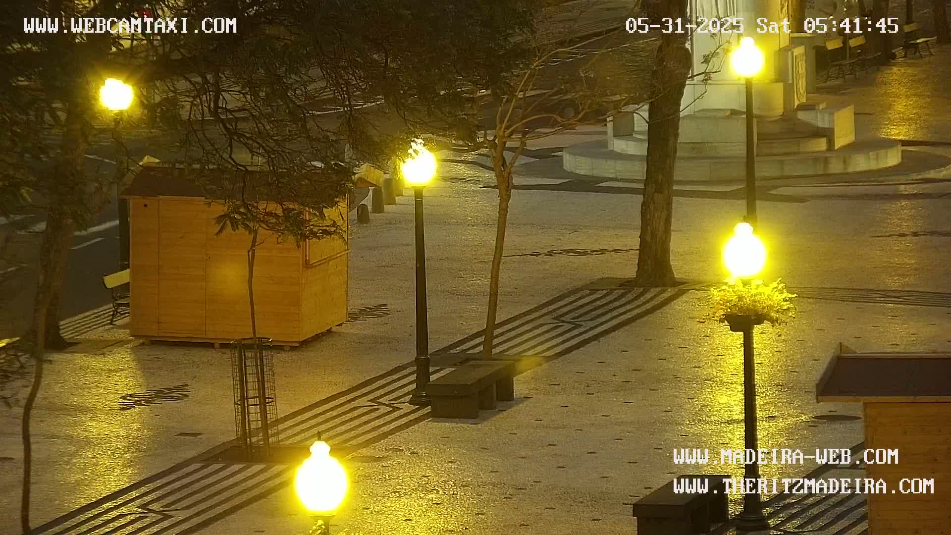 A nighttime, outdoor scene shows a paved plaza illuminated by several bright lamps, with a small wooden structure and a bench visible.