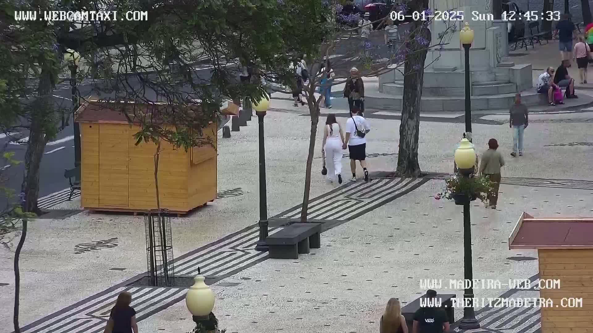 A paved pedestrian area with several people walking, a small wooden kiosk, and street lamps, on an overcast day.
