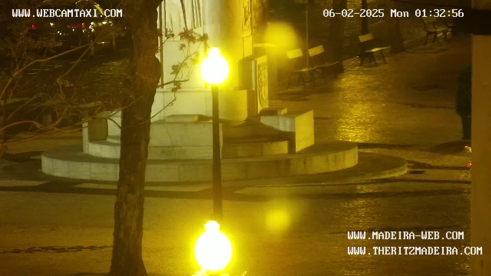 A wet, nighttime scene shows a multi-tiered stone structure illuminated by bright yellow streetlights, with a tree and a few empty benches visible in the background.