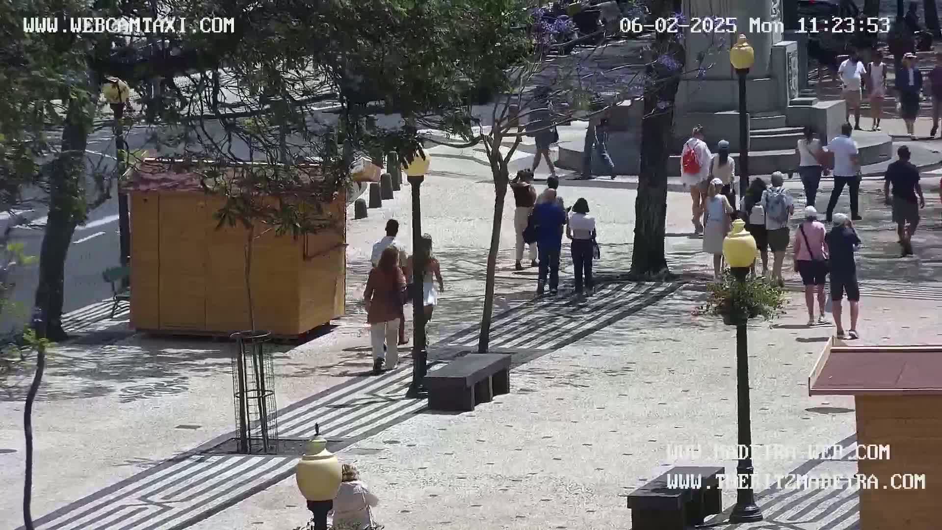 A sunny outdoor plaza features several people walking on a paved area with striped patterns, passing by a small wooden kiosk and benches under leafy trees.