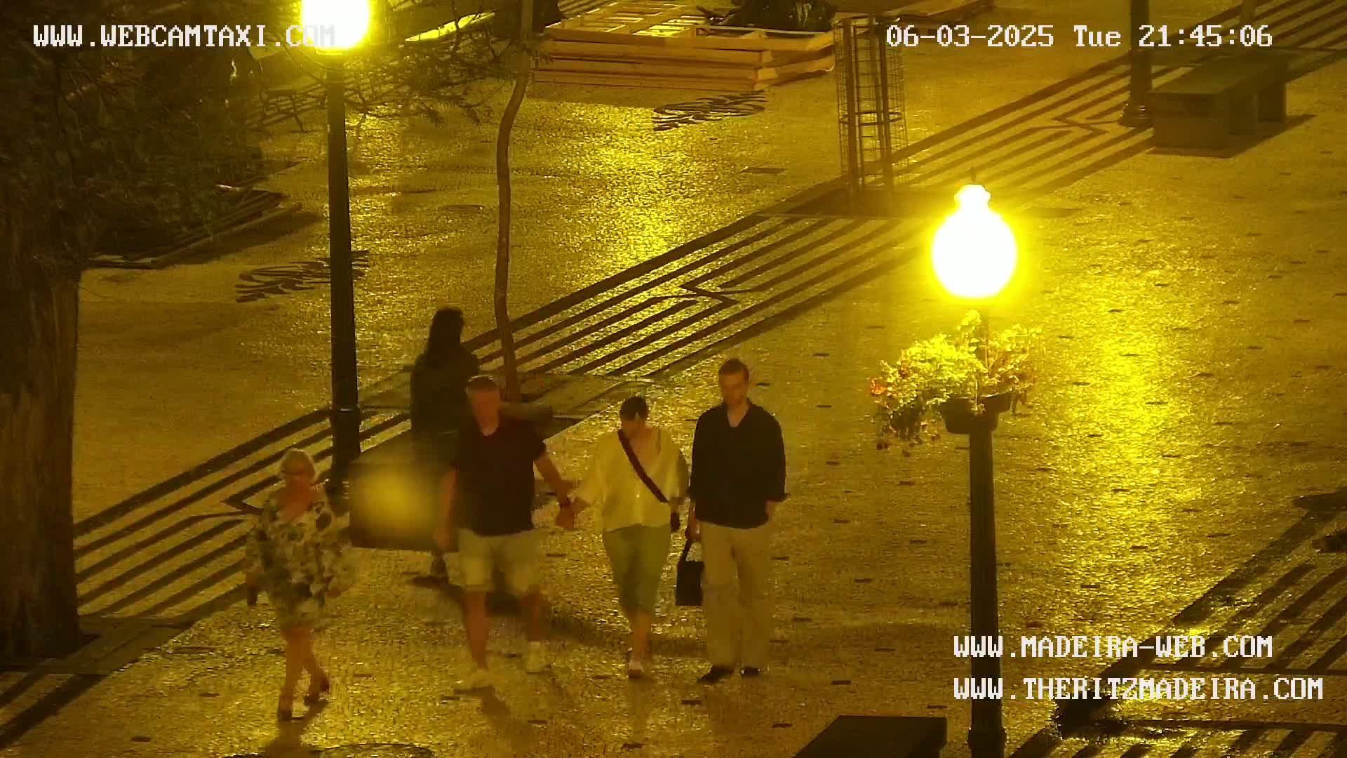 A wet, nighttime cobblestone street is illuminated by a streetlamp, where several people walk in pairs.