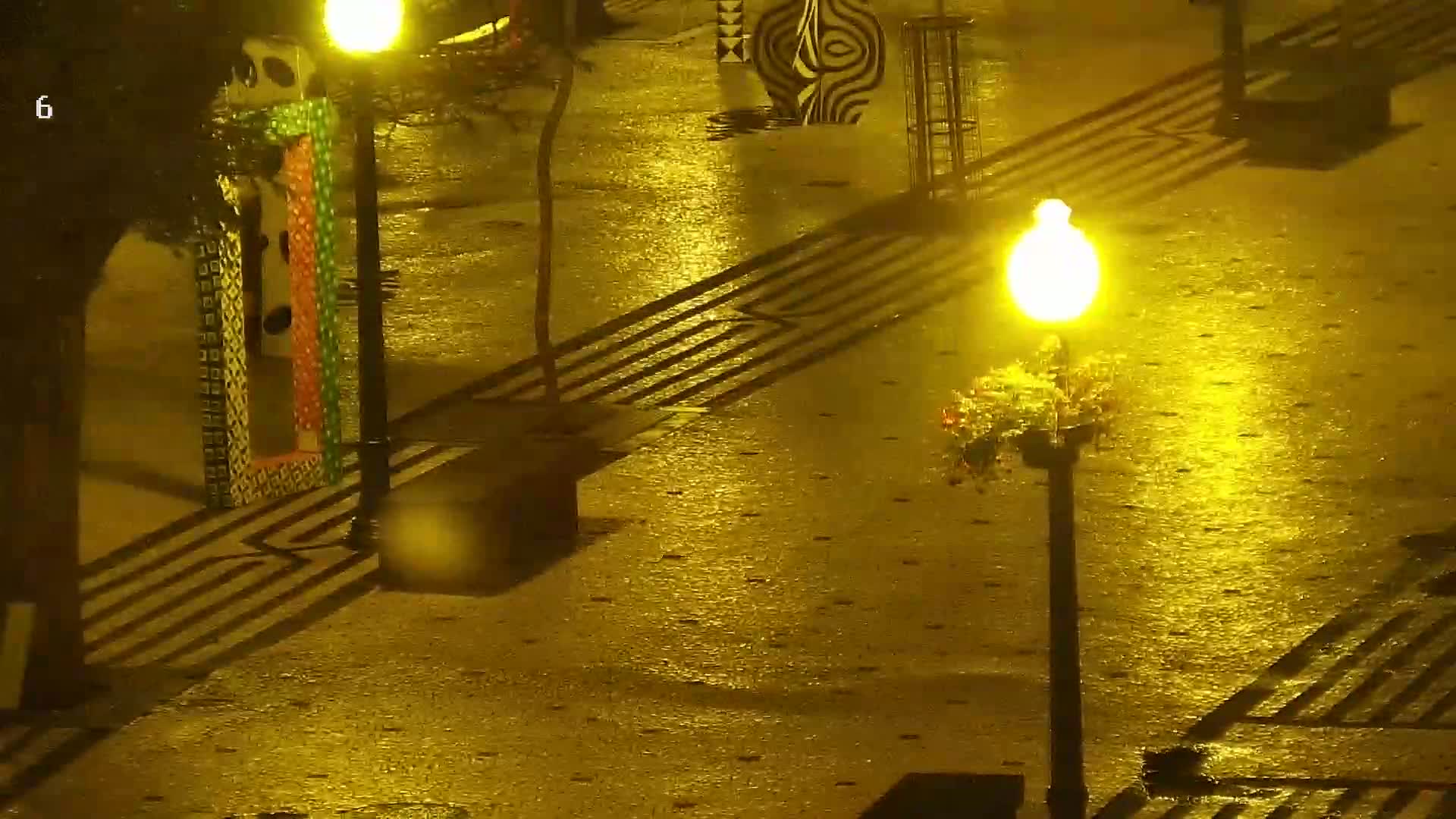 A rain-slicked paved area at night is illuminated by several bright street lamps, casting shadows from nearby decorative structures and railings.
