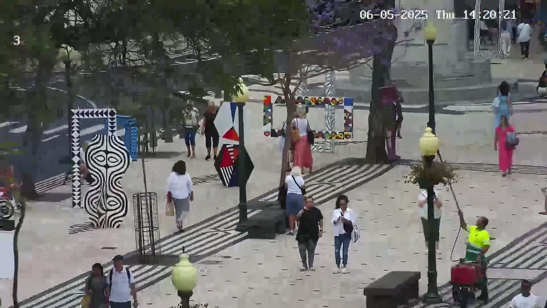 A paved outdoor plaza with geometric art installations and several people walking around on a partly cloudy day.