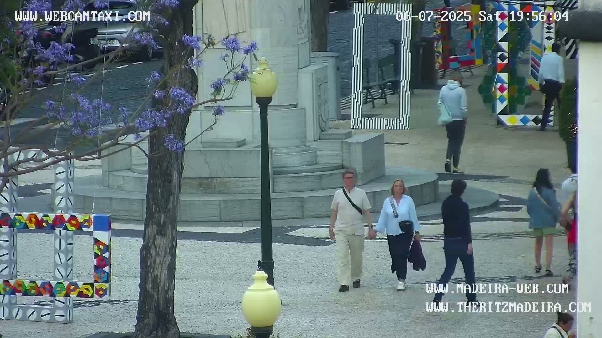 A sunny outdoor plaza scene shows several people walking past colorful, geometric art installations and a stone monument, with a jacaranda tree in bloom.