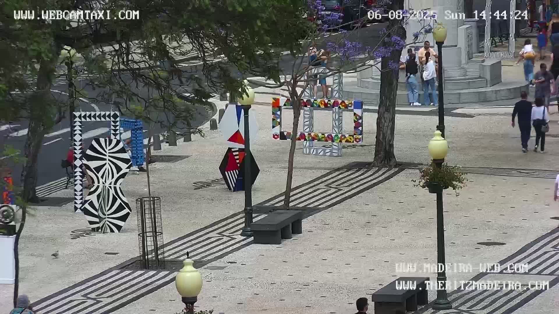 A paved plaza with several modern, colorful art installations and people walking around on a sunny day.