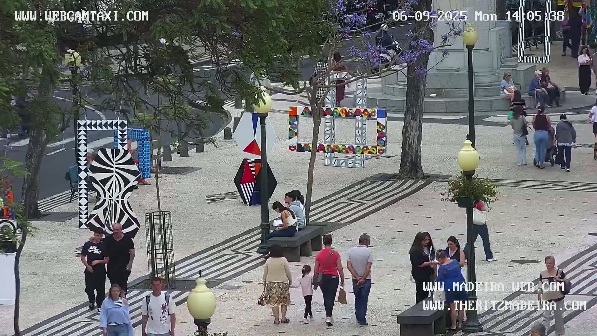 A sunny outdoor plaza features several people walking and sitting, along with various abstract geometric art installations displayed on the paved ground.
