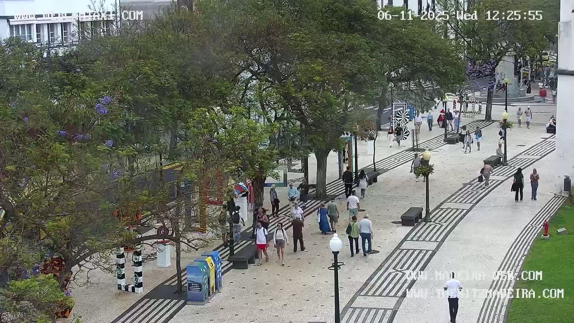A paved pedestrian area with geometric patterns is filled with people walking under a partly cloudy sky, surrounded by lush green trees and various colorful street art installations.