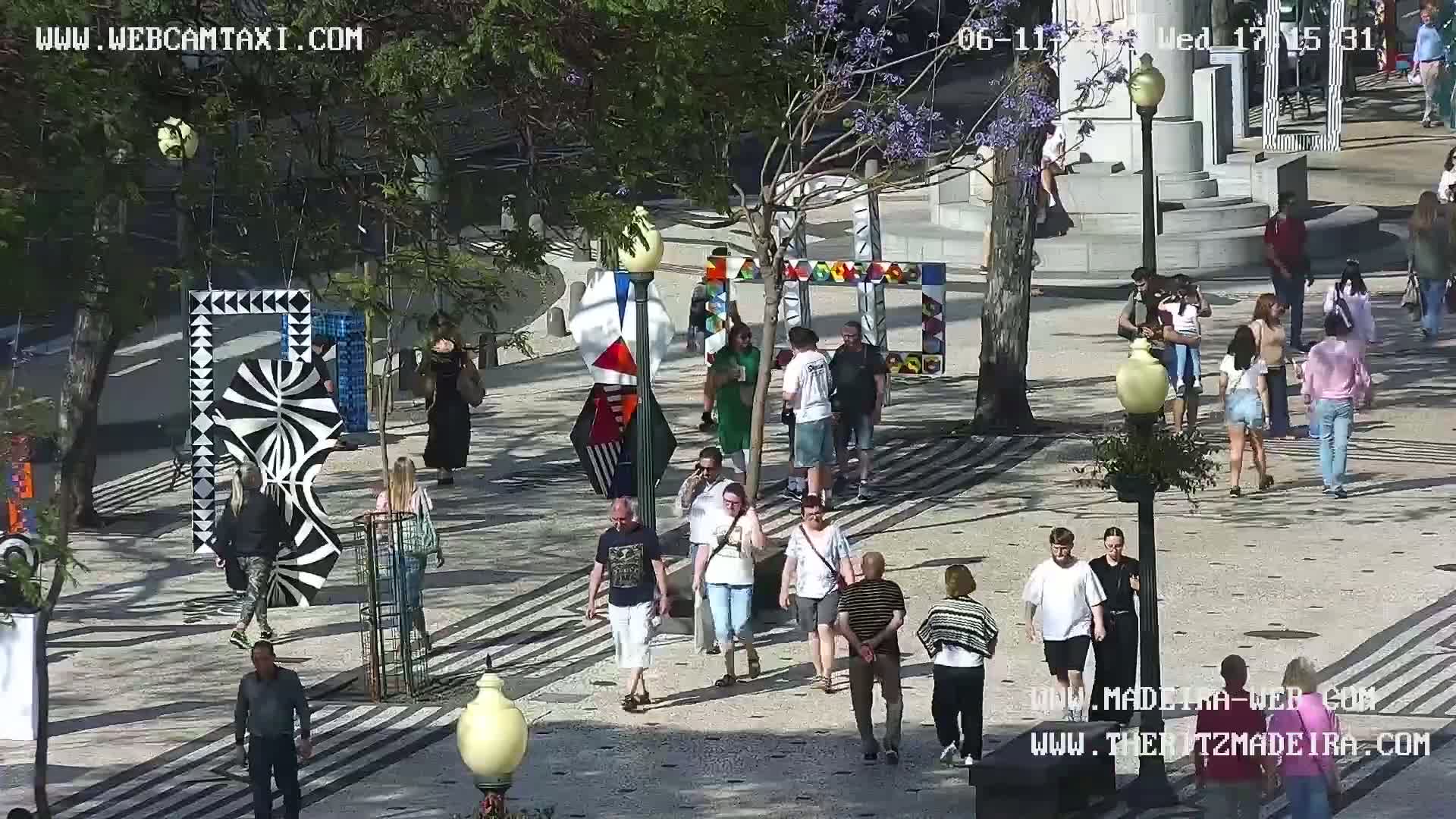 A sunny outdoor plaza features numerous pedestrians strolling among various colorful, geometric art installations.