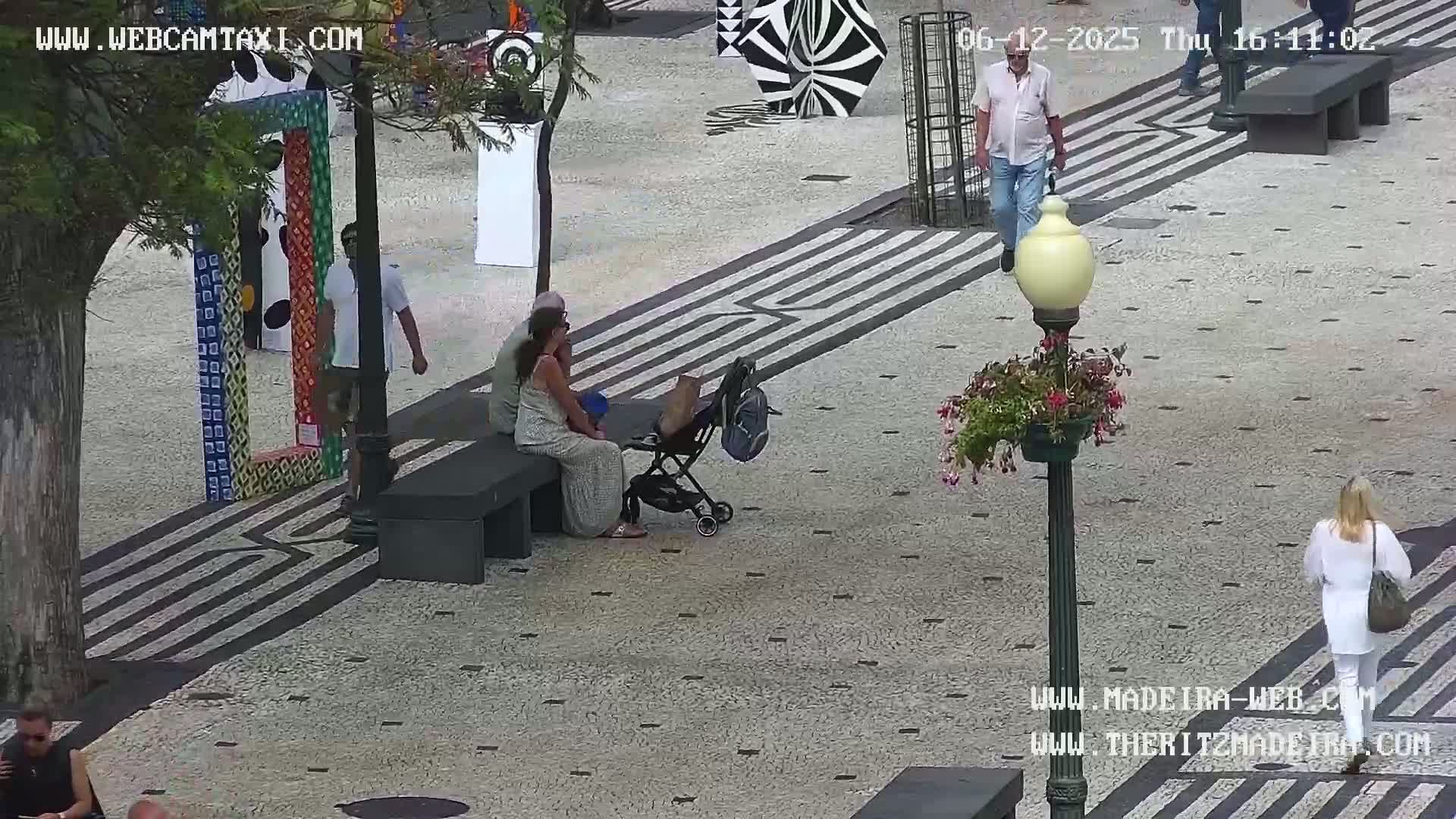 A paved outdoor plaza with several people walking and sitting, some colorful artwork, and a stroller.