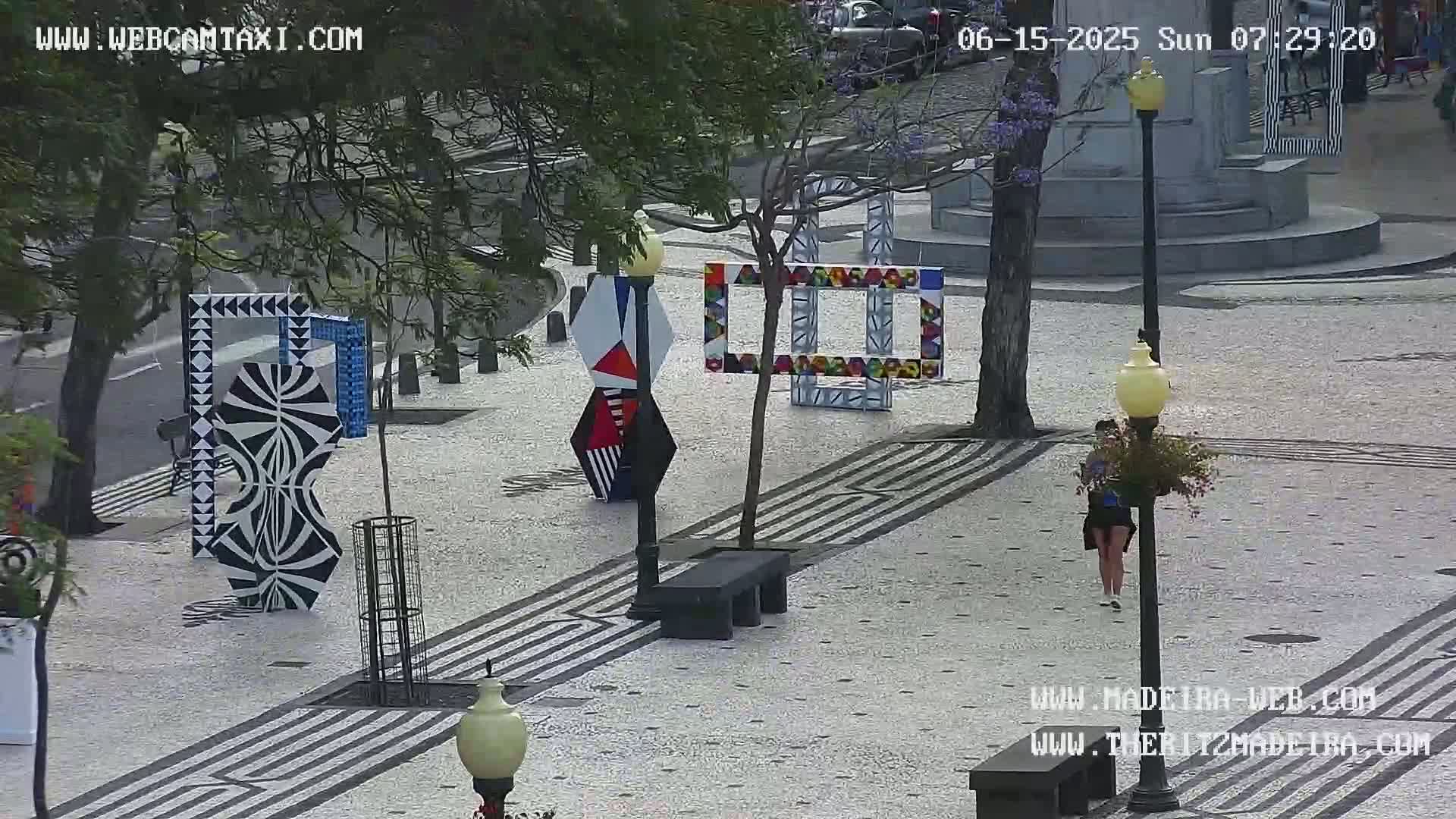 A paved plaza with abstract, colorful geometric art installations, a few benches, lampposts, and a person walking.