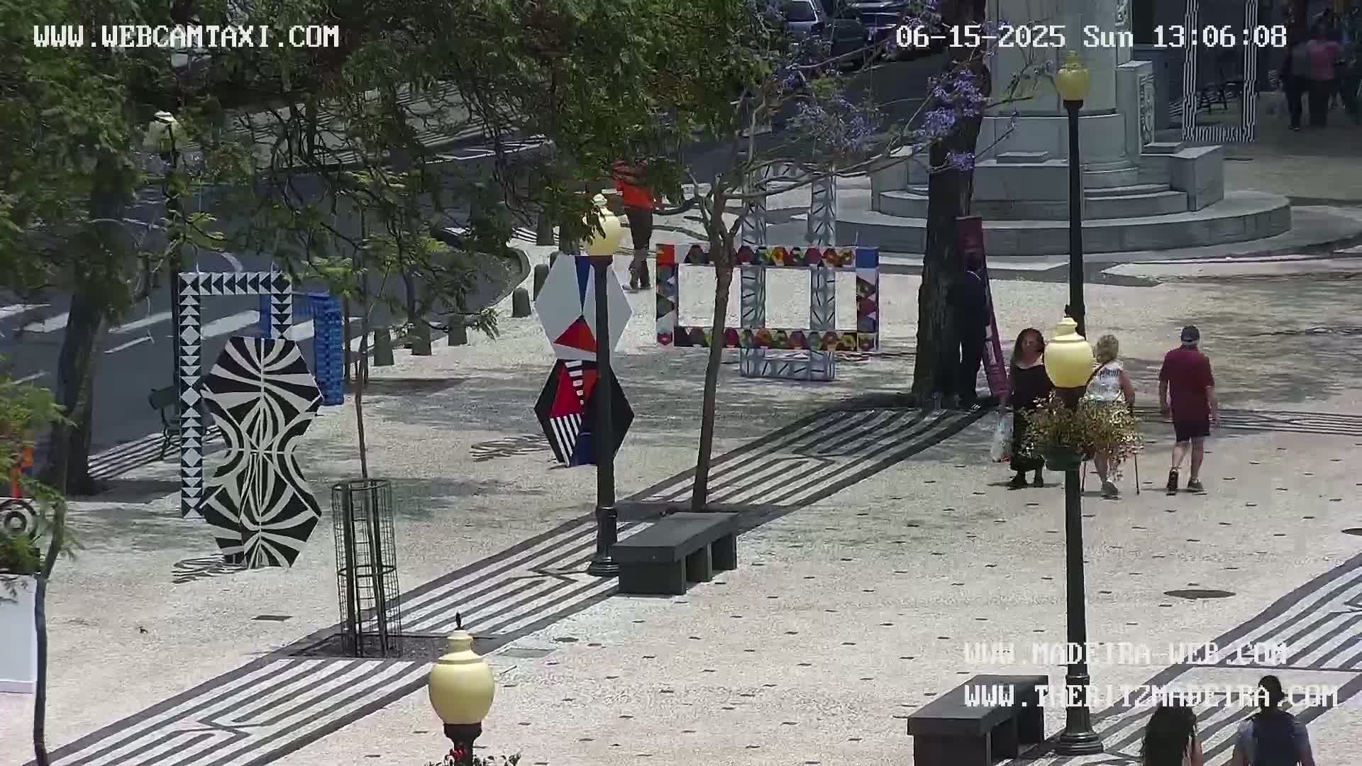 A sunny outdoor plaza features several colorful, geometric art installations and a few pedestrians walking on a patterned stone surface.
