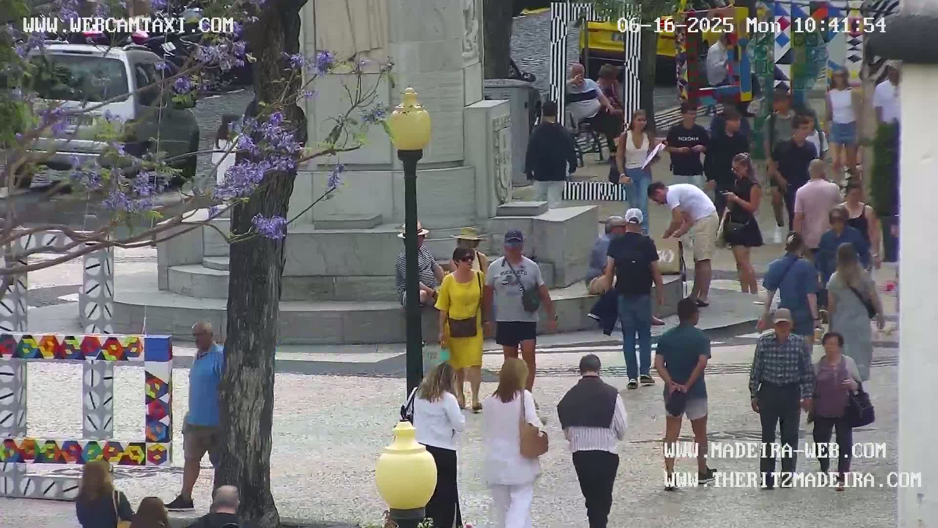 A sunny outdoor plaza scene shows numerous people walking around a monument and colorful artwork, with a tree displaying purple blossoms in the foreground.