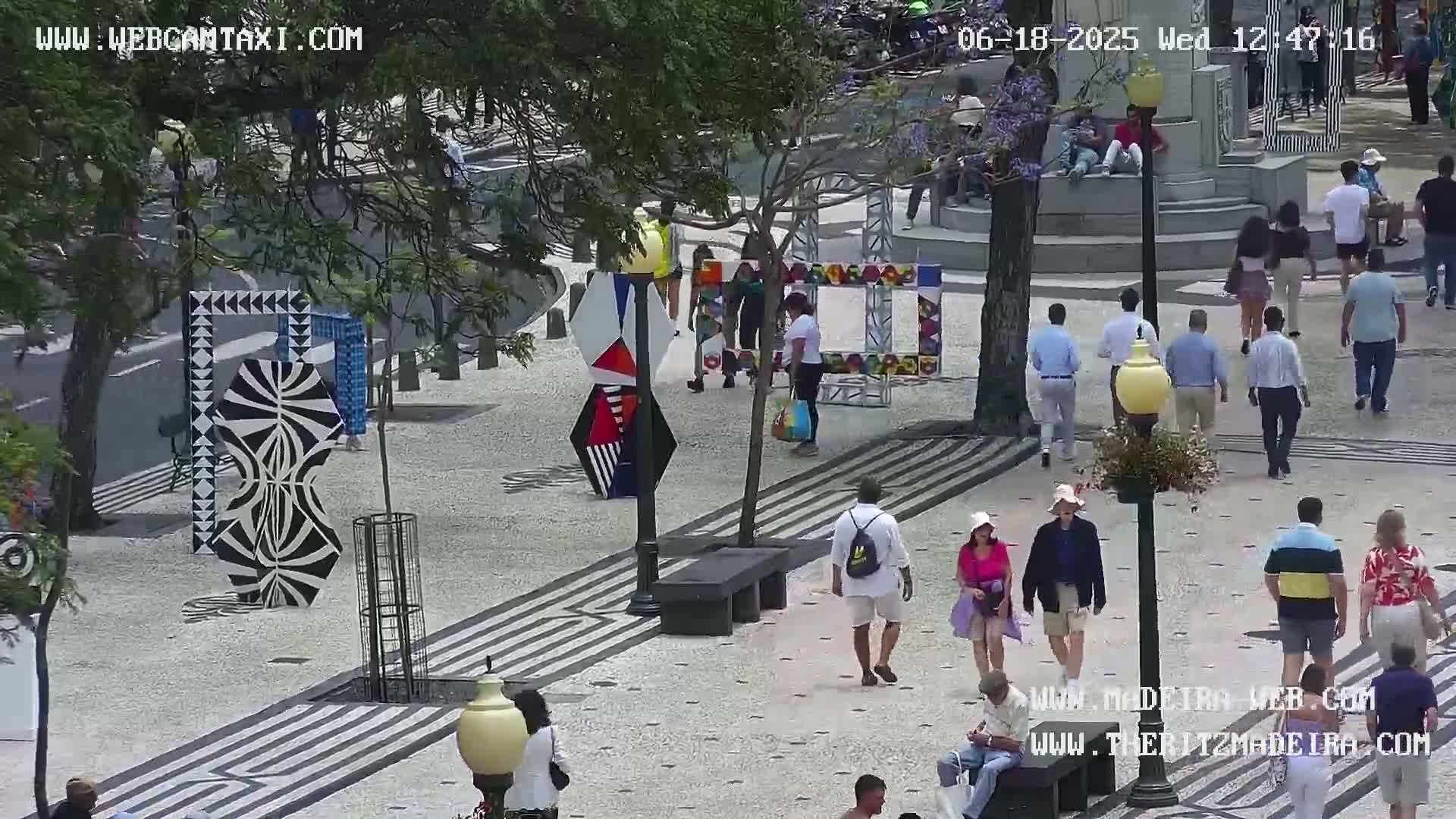 A sunny outdoor plaza features pedestrians walking among colorful abstract sculptures and patterned pavement.
