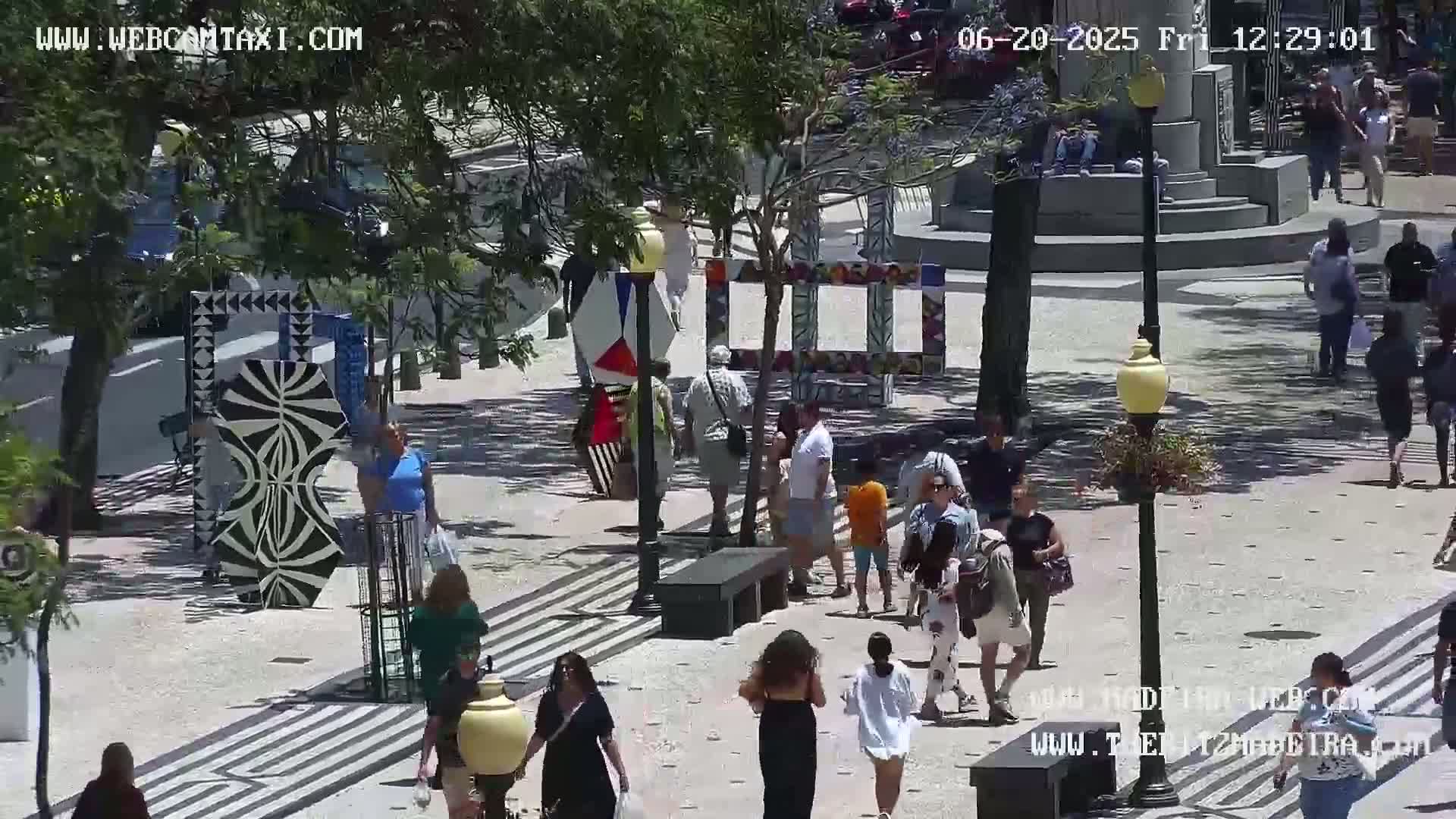 A sunny outdoor plaza features several people walking among modern art installations and street lamps.