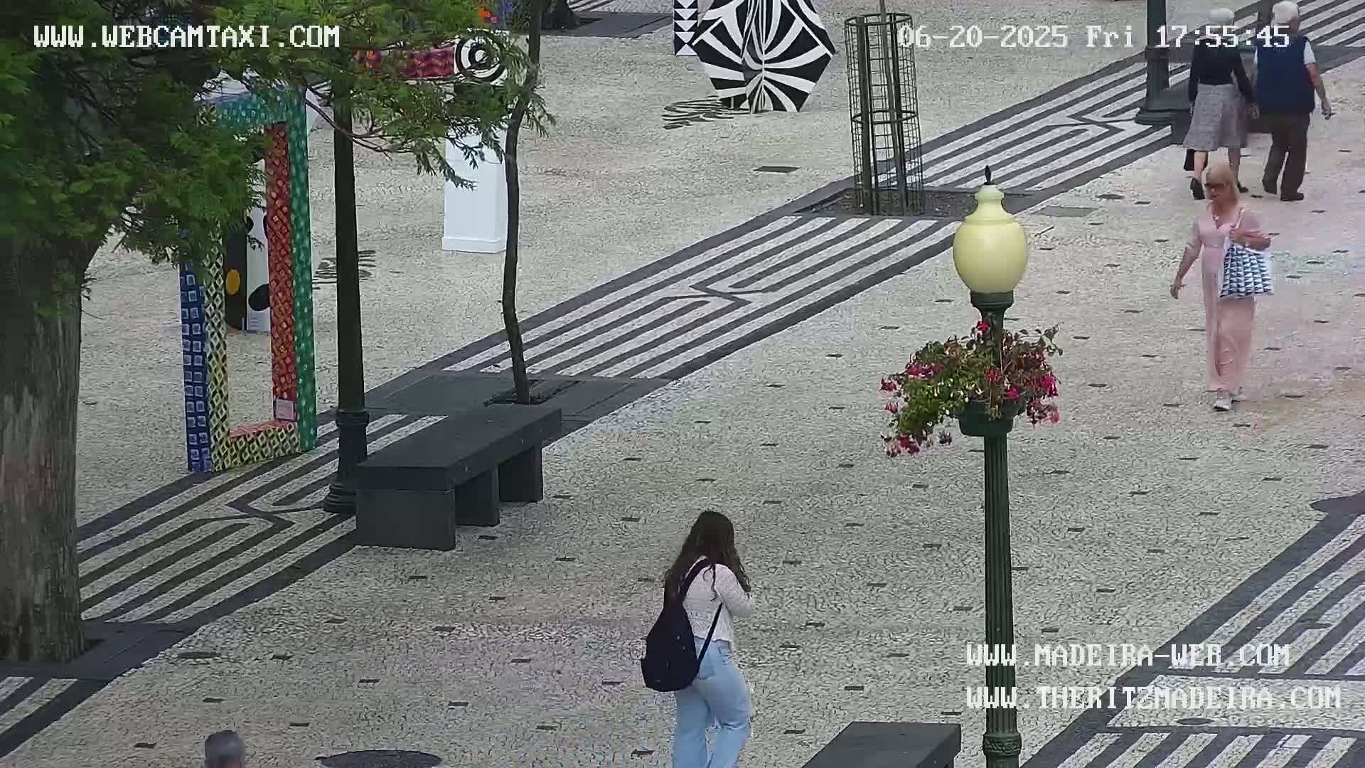 A paved outdoor plaza with several pedestrians, a bench, colorful art installations, and a lamppost with flowers.