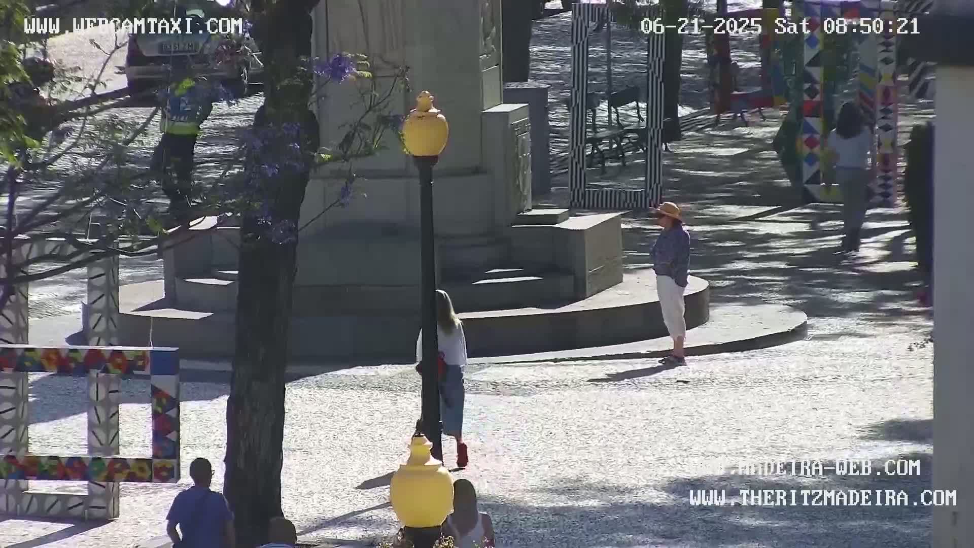 A sunny outdoor plaza features several people walking past a stone monument and colorful artistic structures.