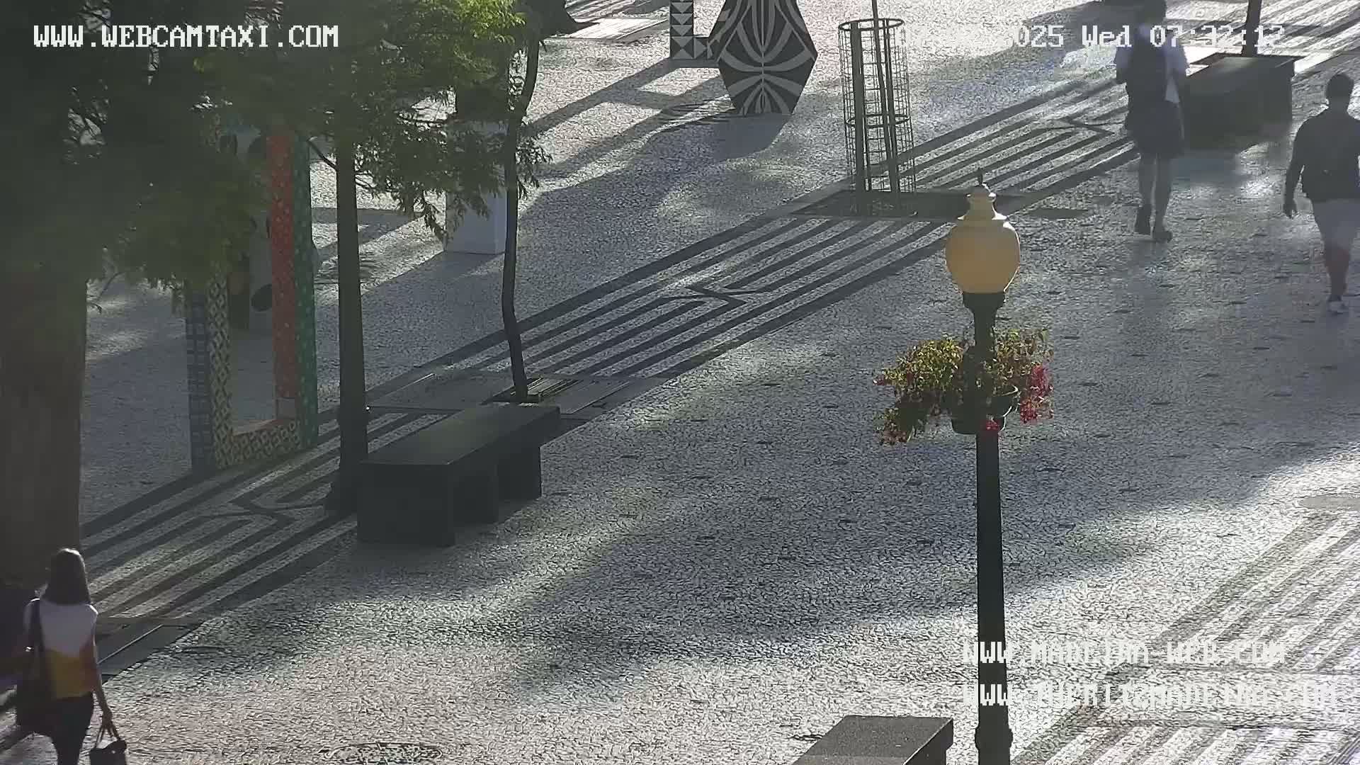 A paved outdoor area with patterned walkways, benches, decorative structures, and lampposts, with people walking by in the sunny weather.
