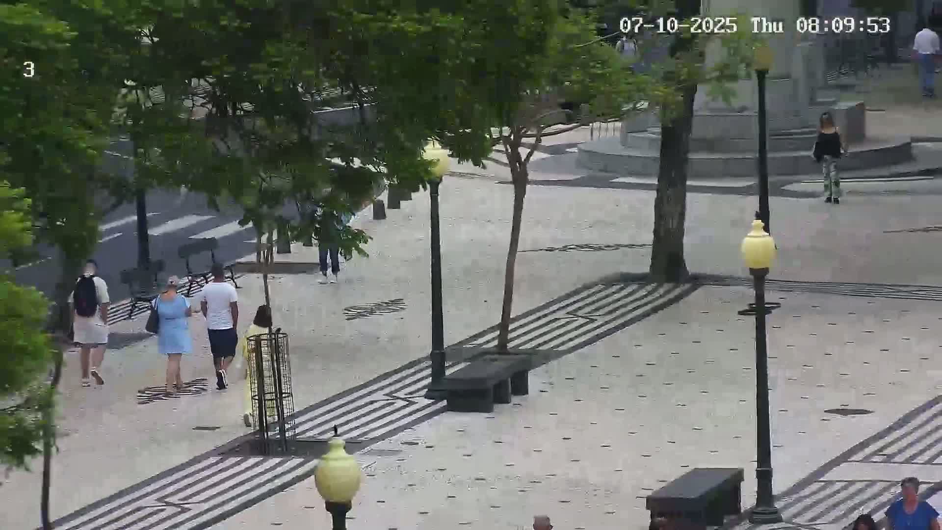 A paved pedestrian area with several people walking, lined with trees and lampposts, under a partly cloudy sky.