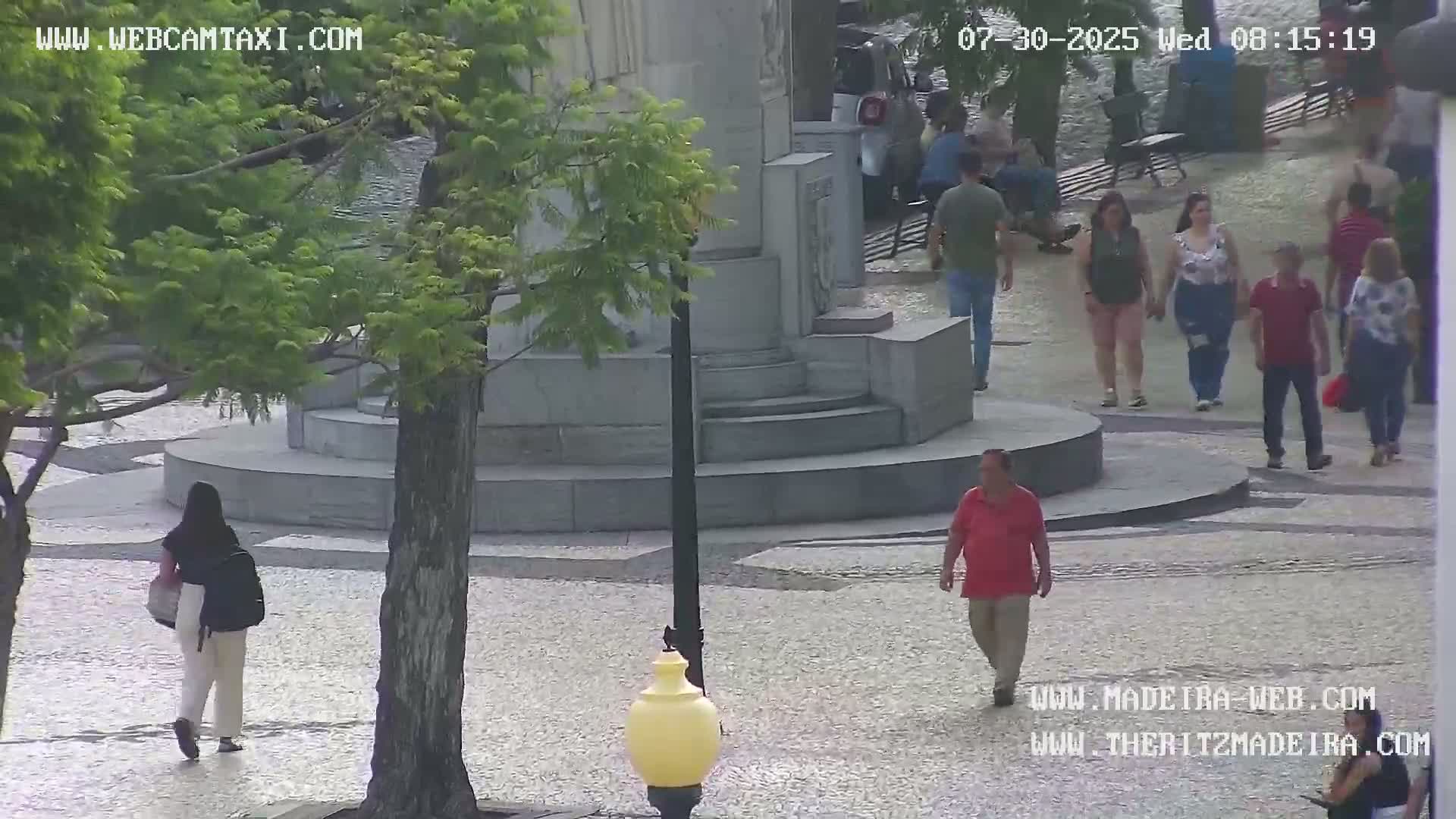 A paved plaza with several people walking around a stone monument, under a partly sunny sky.