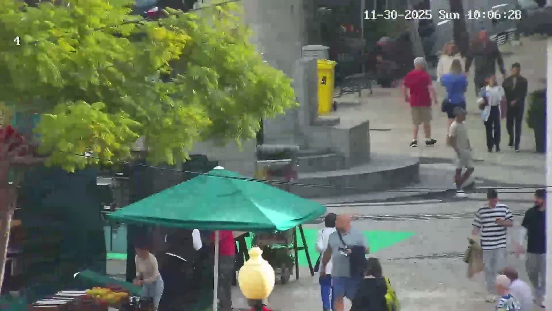 A bustling outdoor scene shows numerous people walking on a paved area near a green-canopied market stall, under clear, sunny daytime conditions.