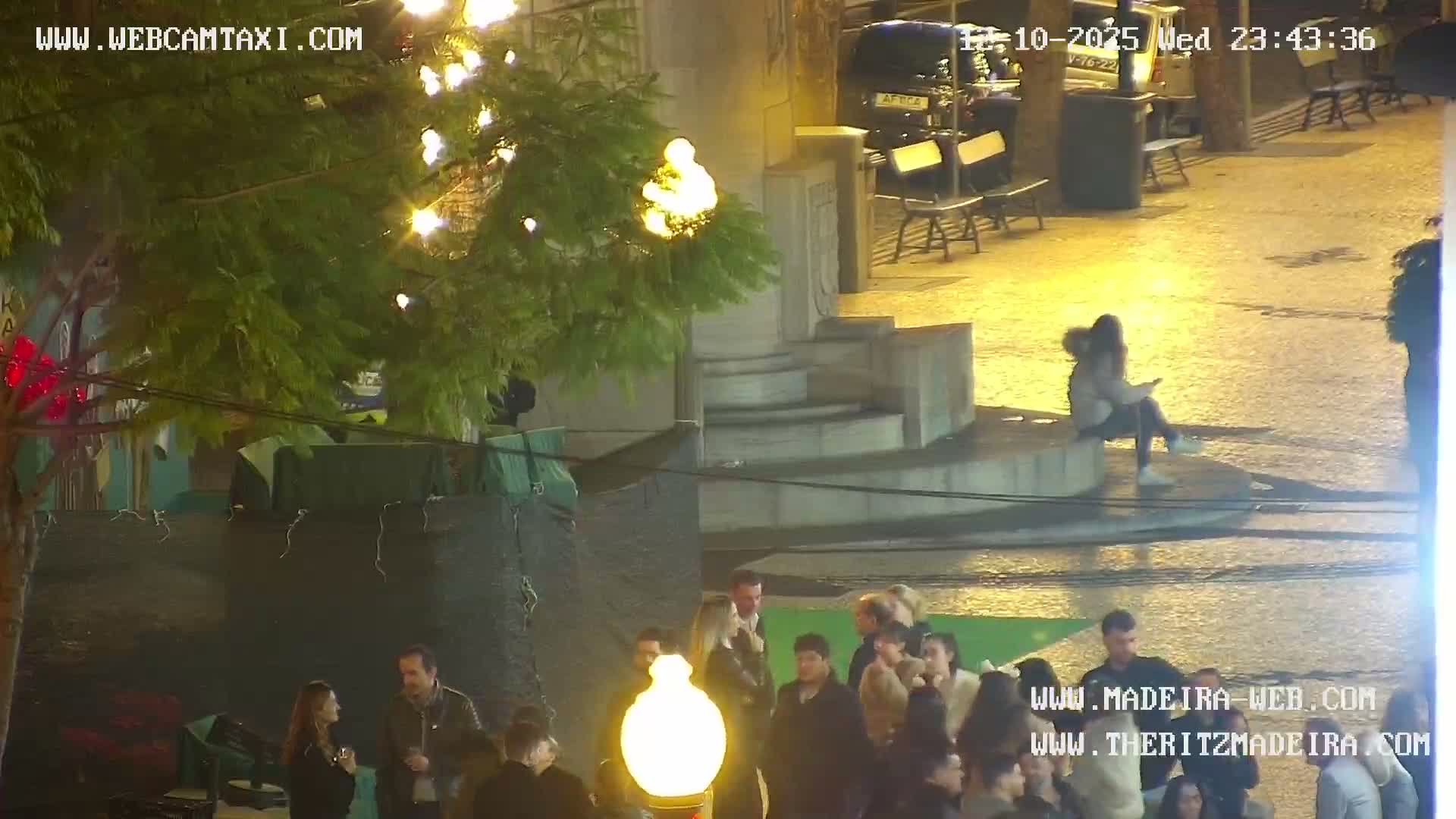 On a clear but damp evening, a bustling outdoor scene unfolds with several groups of people gathering on a wet paved area, illuminated by bright artificial lights that highlight trees and stone structures.