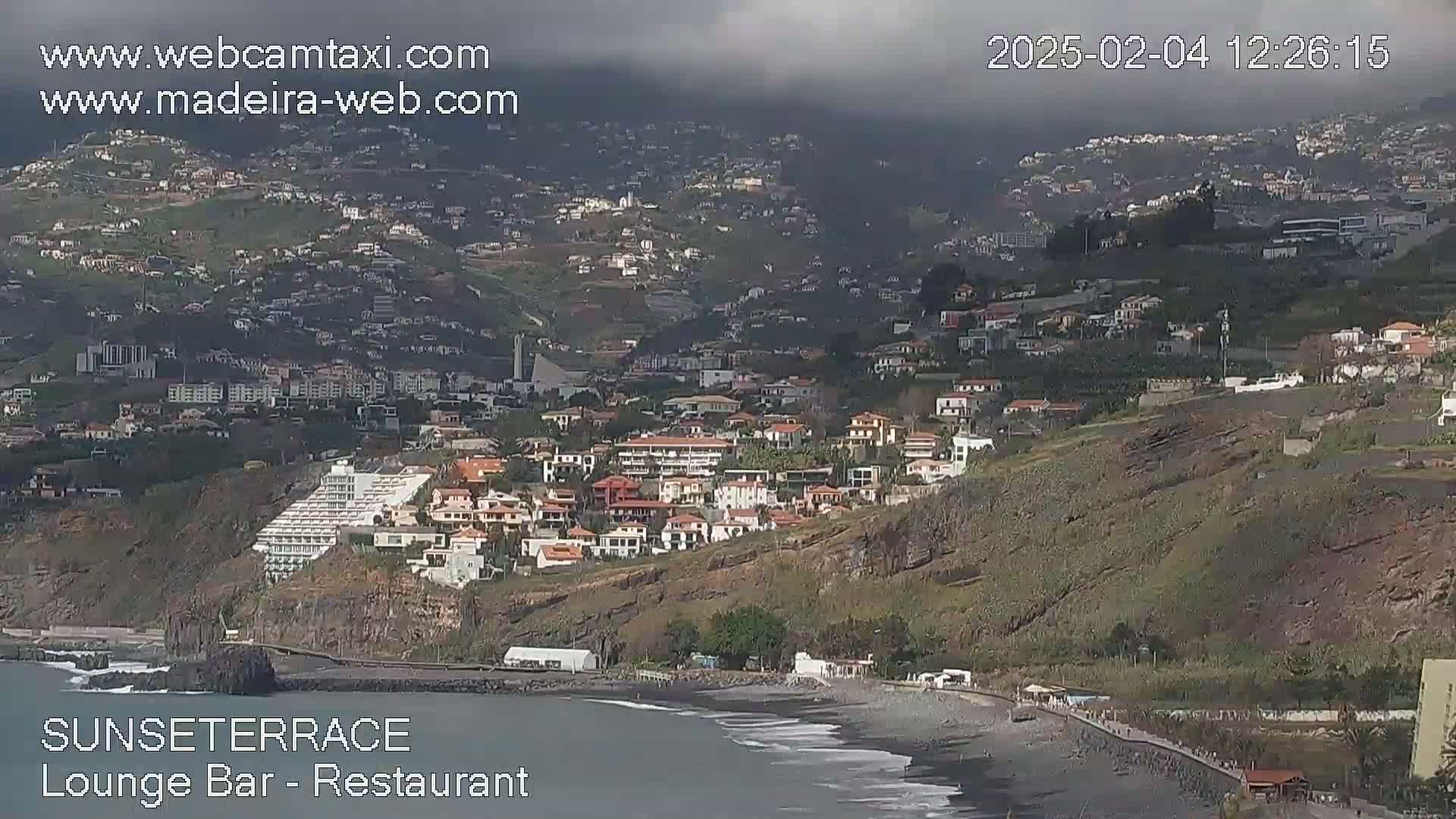 A coastal town with many buildings is visible on a hillside overlooking a dark-sand beach, under an overcast sky.