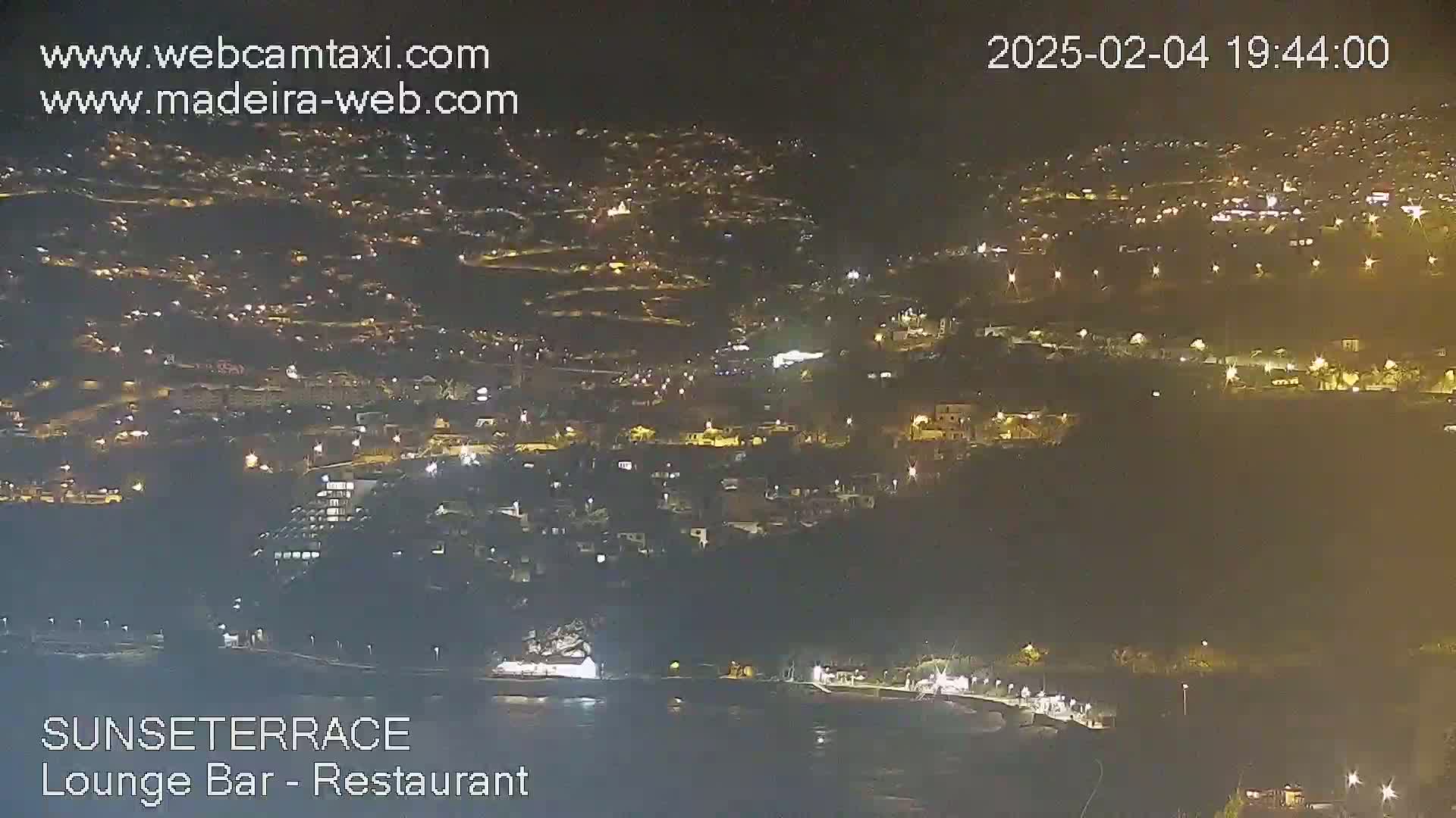 A nighttime, aerial view of a coastal town with numerous lights illuminating buildings and streets, extending down to a dark body of water.