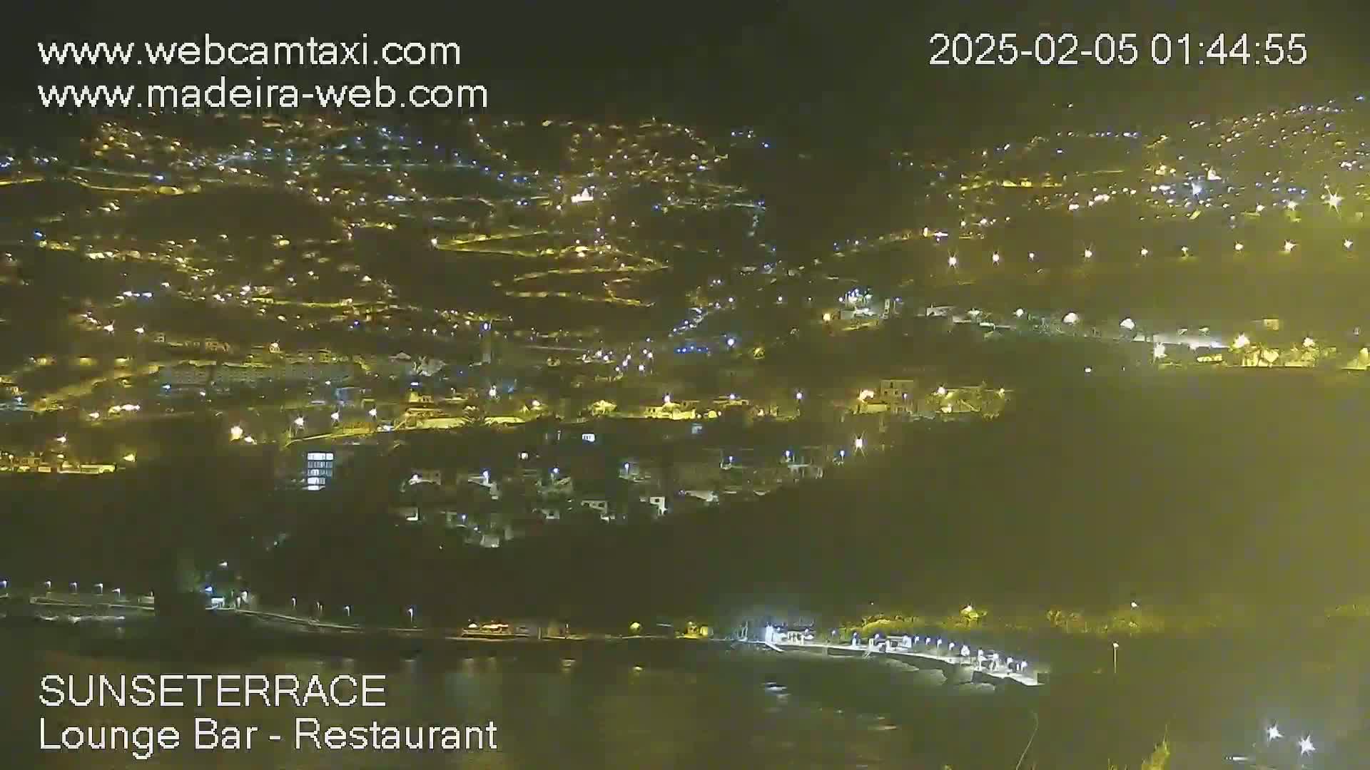 A nighttime, aerial view shows a hillside town with numerous lights illuminating buildings and streets, along a dark body of water.