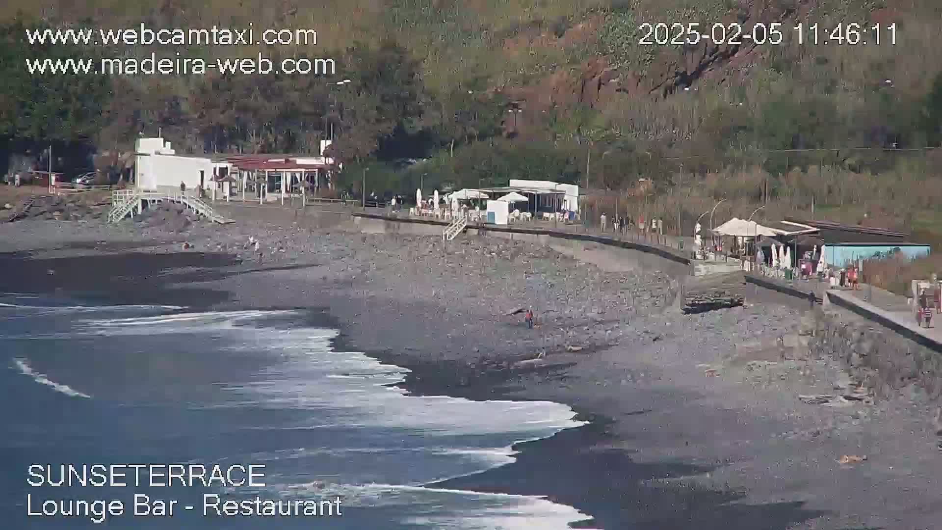 A dark-sand beach with gentle waves laps against the shore near a beachfront restaurant and promenade, under a sunny sky.