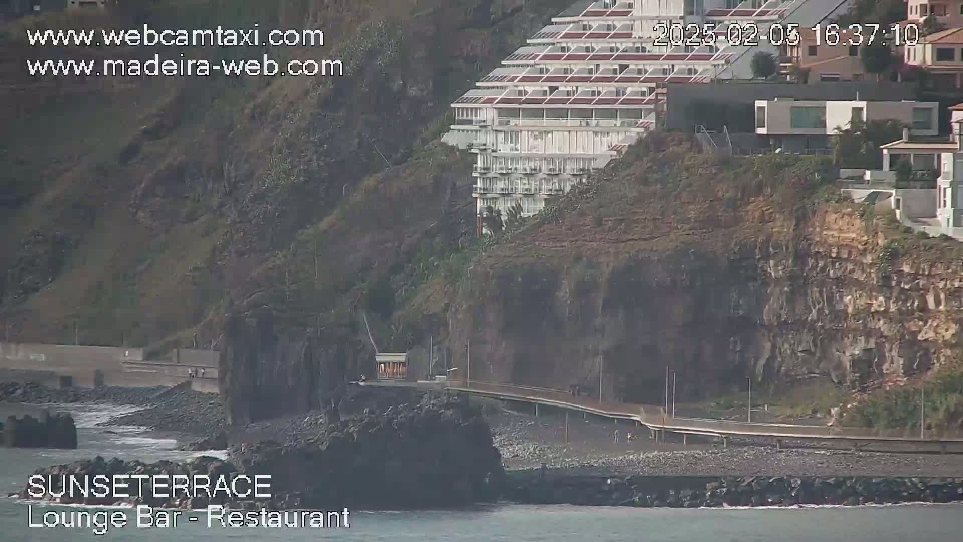 A coastal scene shows a multi-story building perched atop a rocky cliff overlooking a dark-colored beach and calm ocean water.