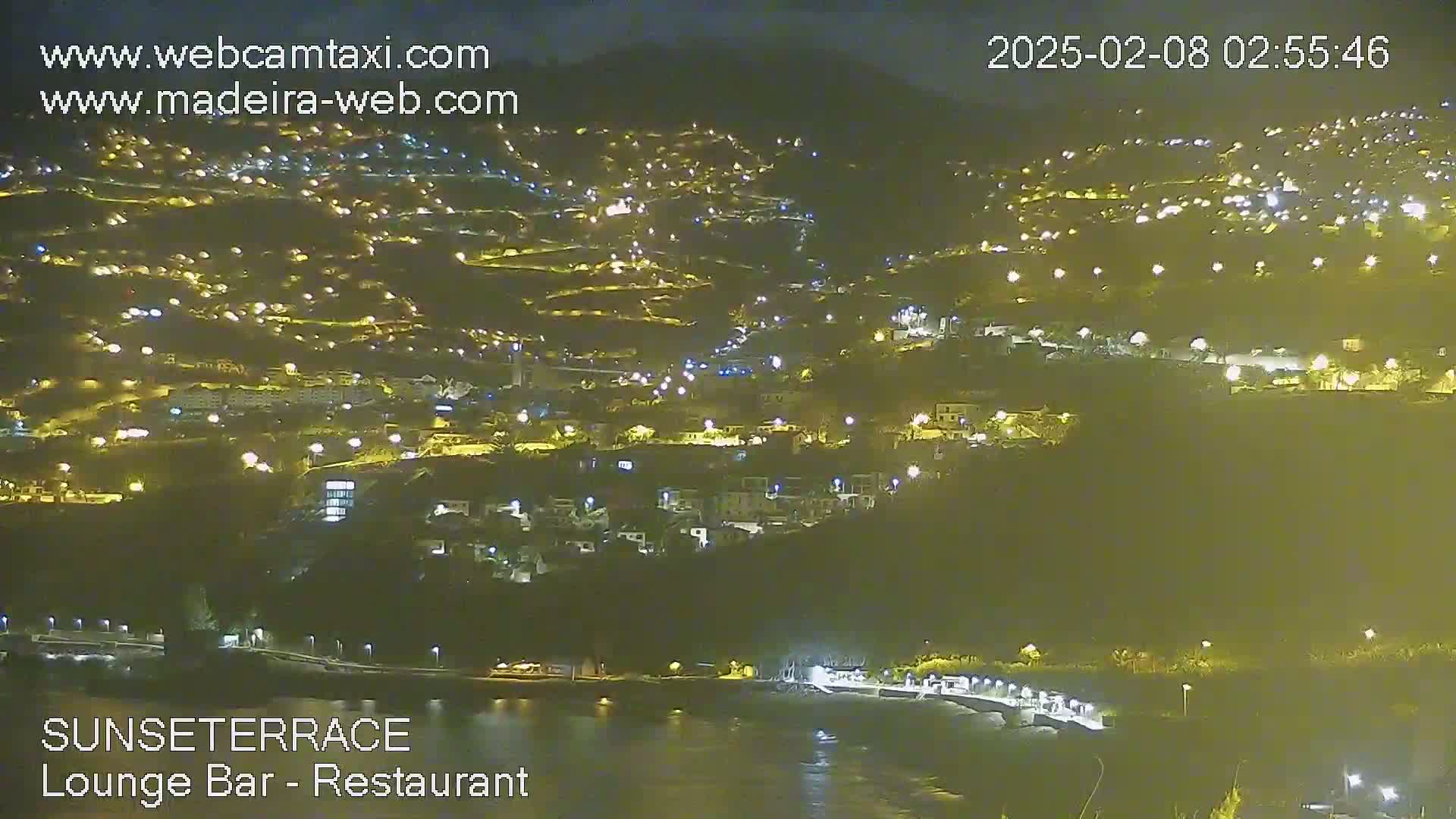 A nighttime, aerial view of a coastal town with many lights illuminating buildings and streets, next to a dark body of water.