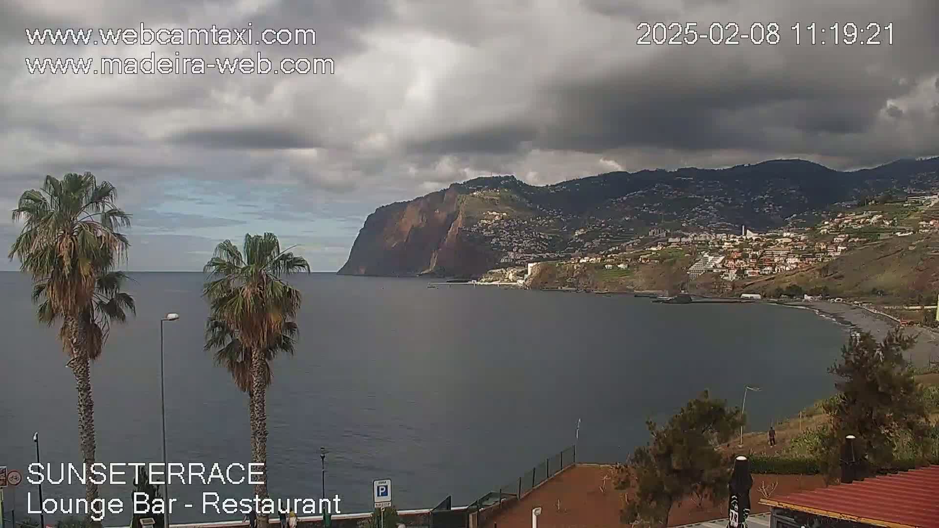 A coastal view shows a dark-grey ocean under a cloudy sky, with a steep, dark-red cliff and a town built into a hillside in the background.