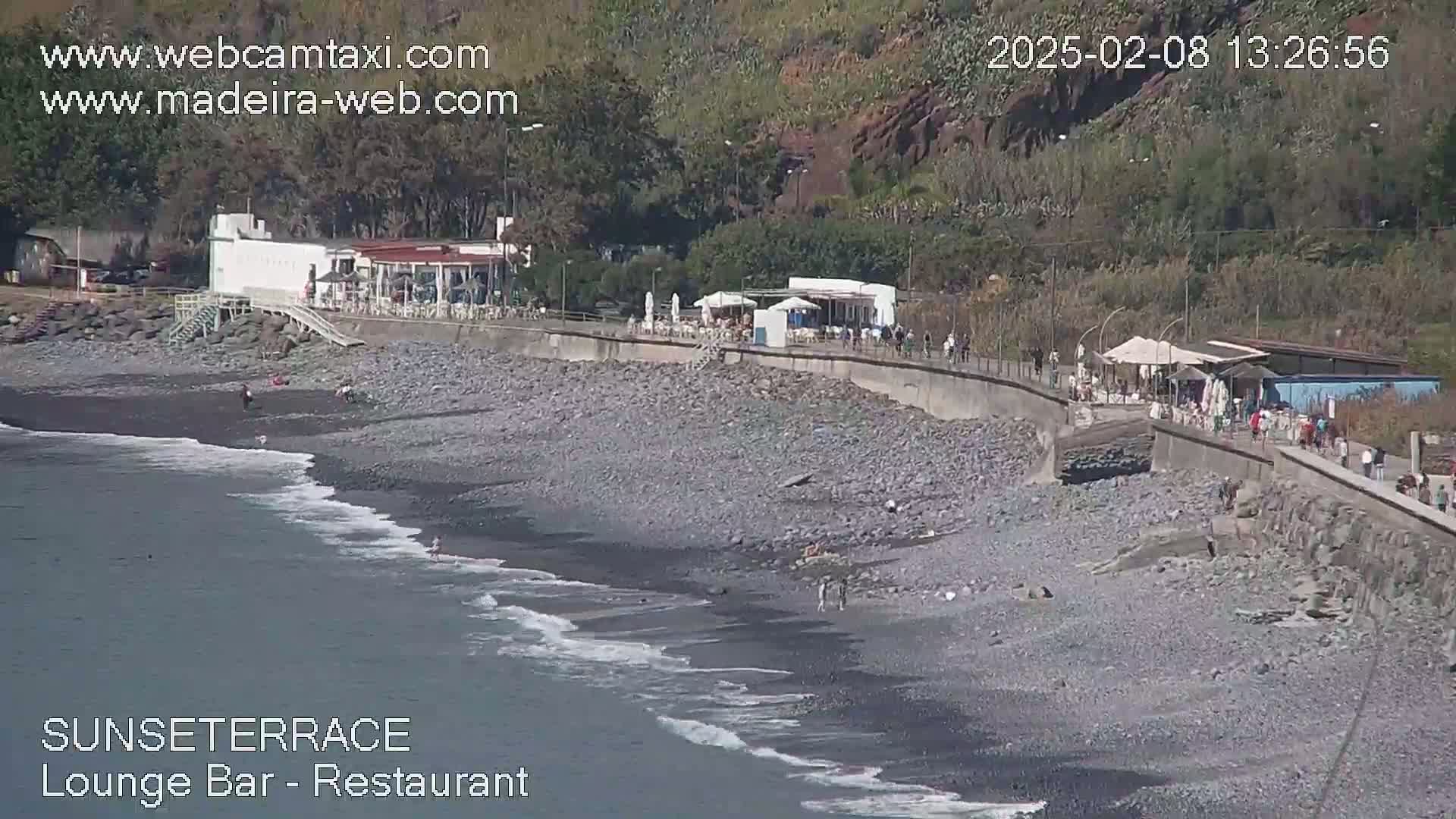 A rocky beach with a restaurant and people walking along a seawall, under sunny conditions with gentle waves.