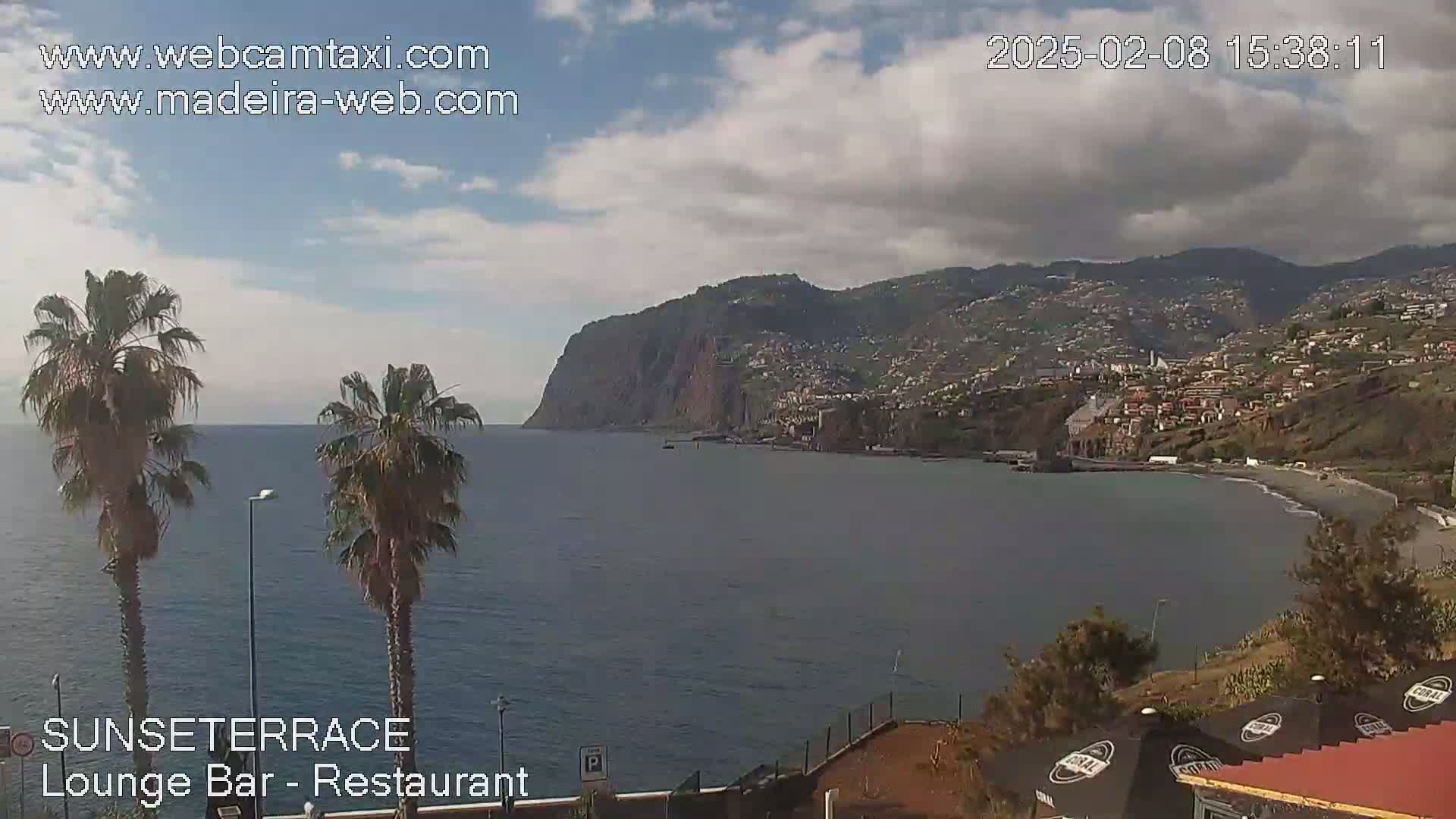 A partly cloudy day reveals a coastal scene with a calm ocean, palm trees in the foreground, and a town nestled against a large cliff in the background.