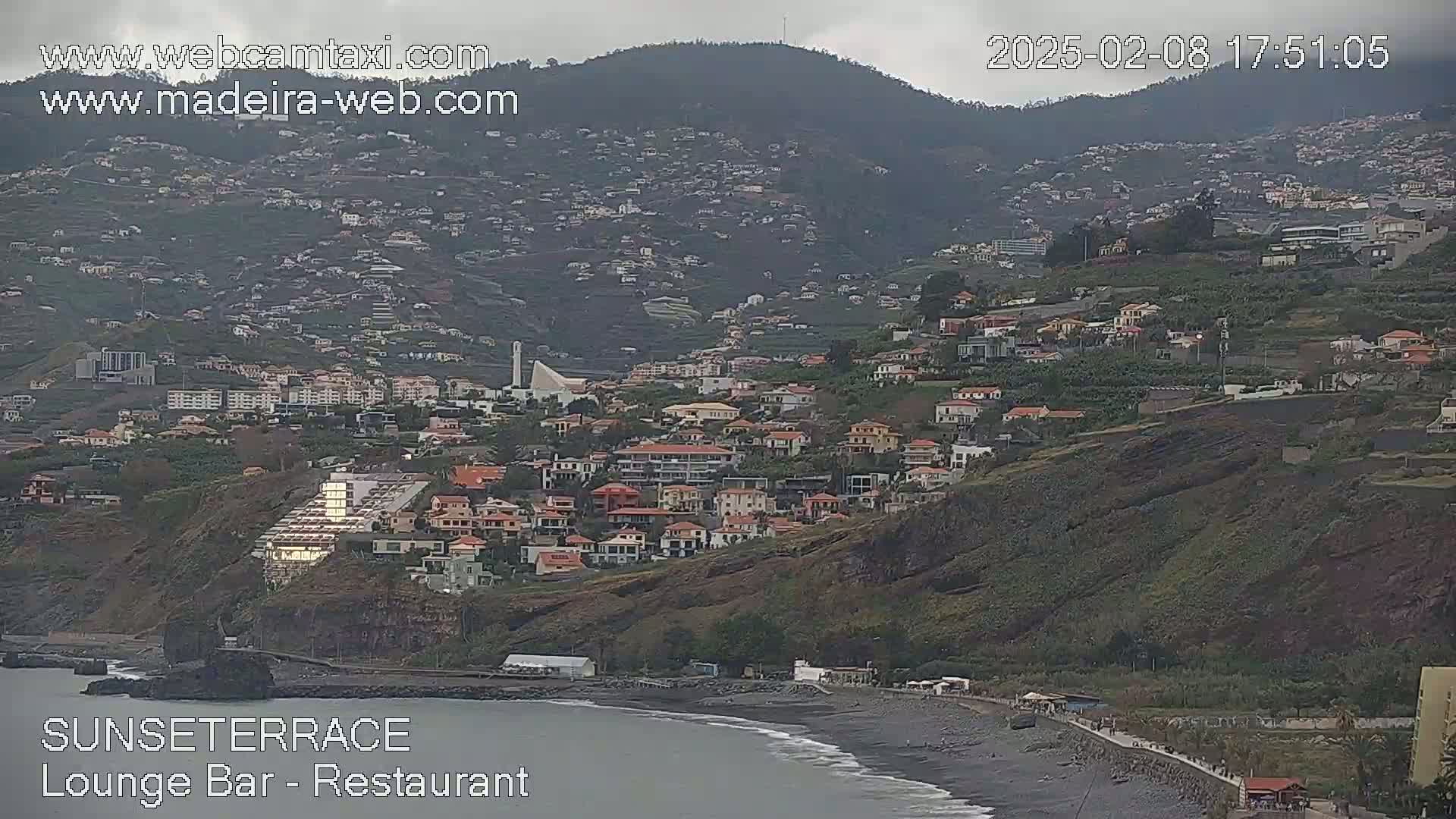 A coastal town nestled on a hillside below a mountain range, under an overcast sky, has a dark-sand beach below it.