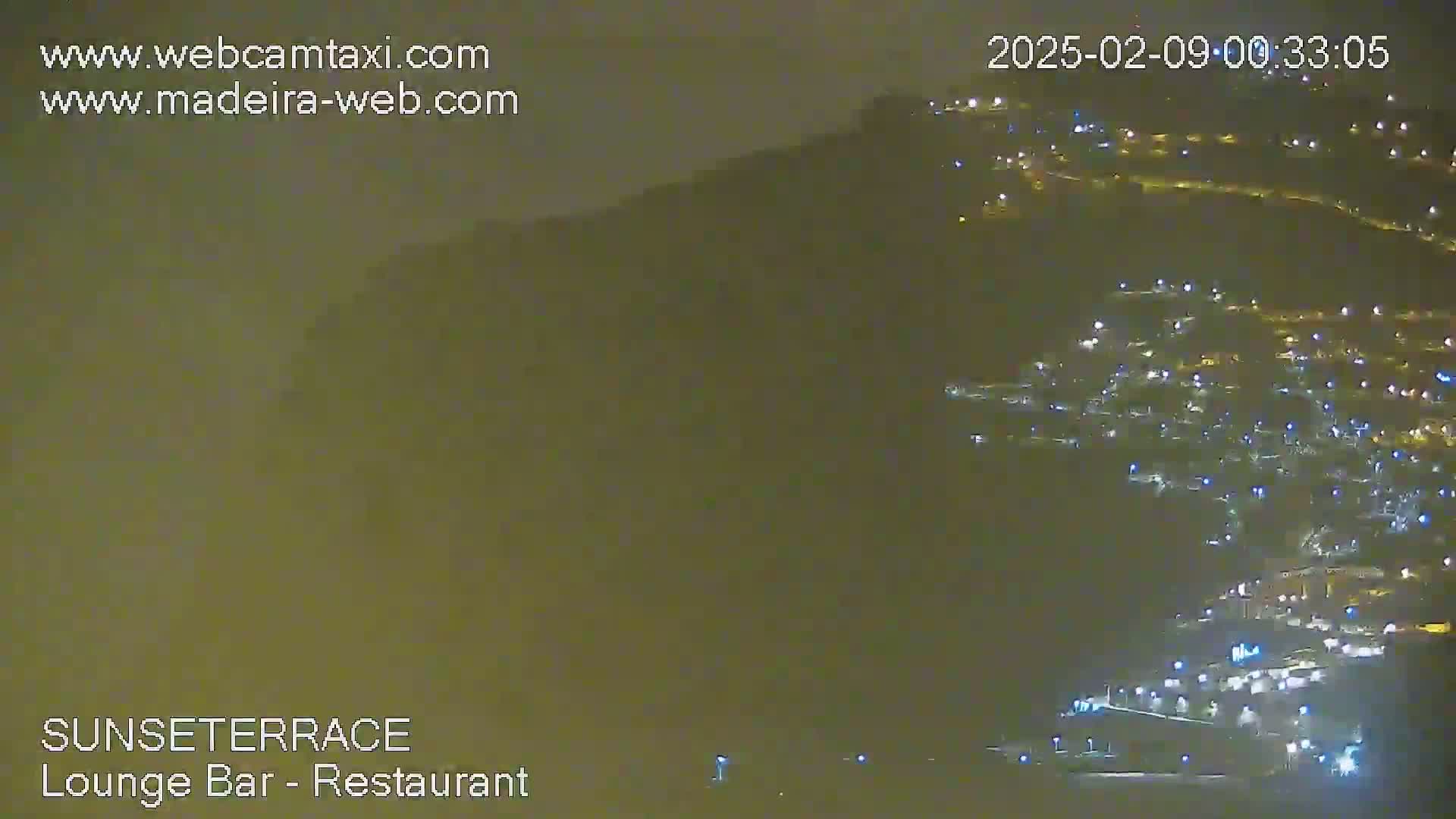 A hazy night scene shows a hillside town illuminated with numerous lights under a dark sky.