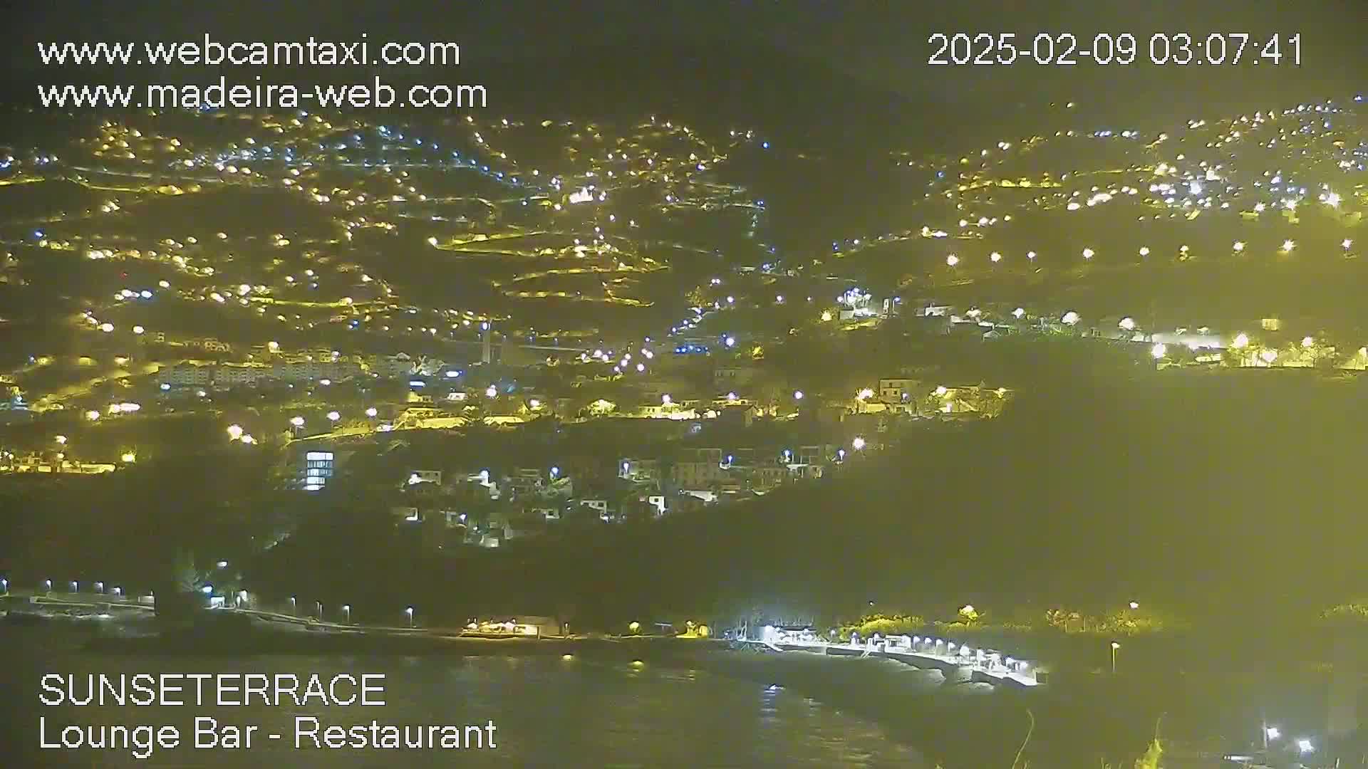 A nighttime view of a hillside town illuminated by numerous lights, overlooking a dark body of water.