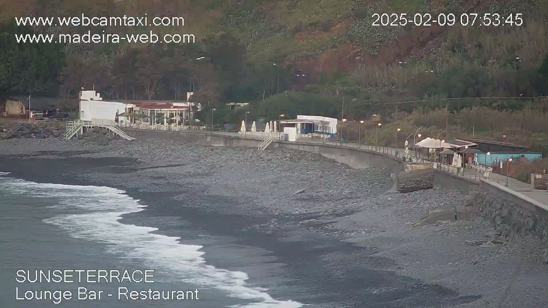 A rocky beach with gentle waves laps at the shore near a beachfront promenade lined with buildings and outdoor seating areas, under an overcast sky.