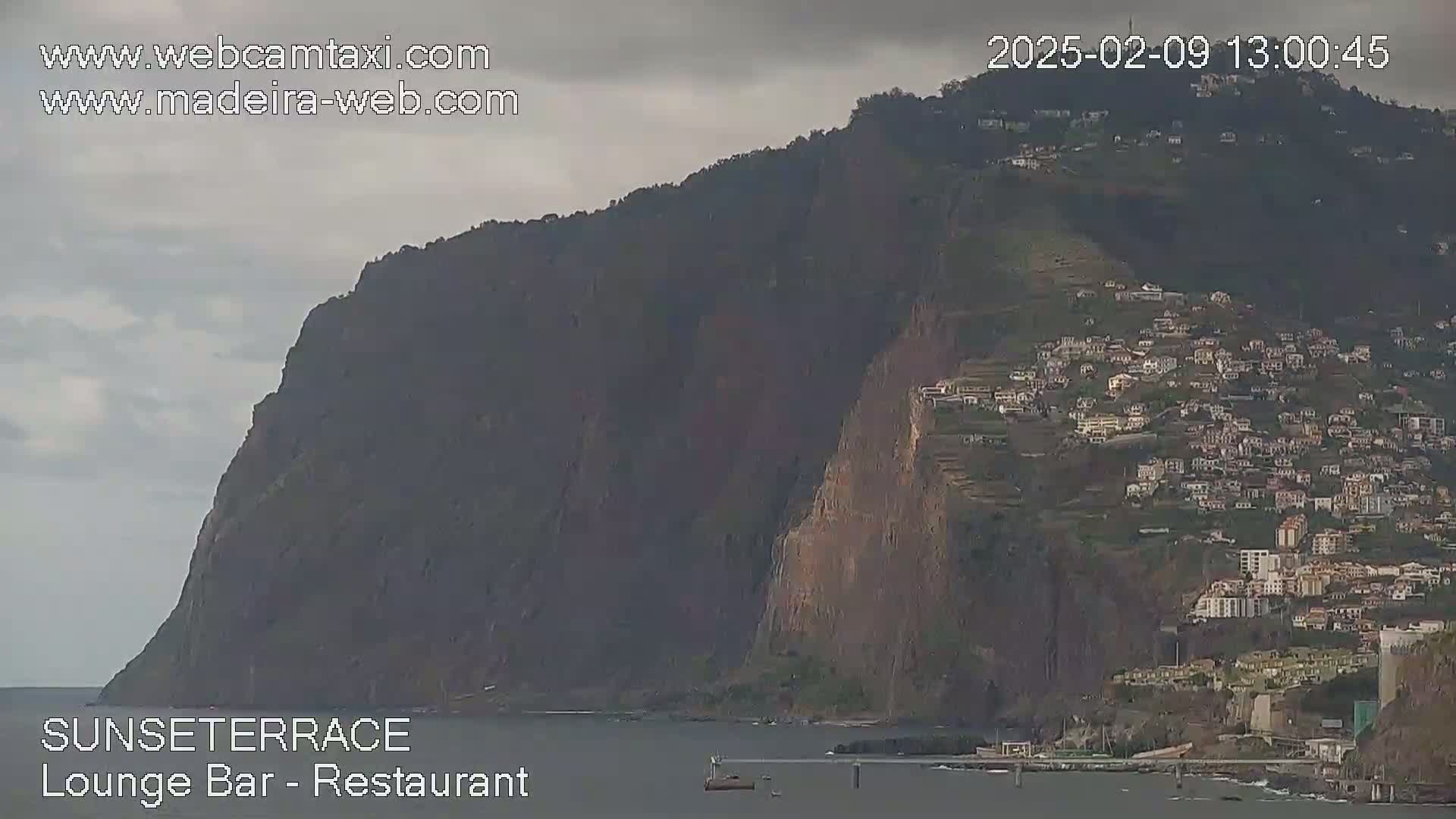 A large, dark cliff overlooks a town nestled on a hillside, all under an overcast sky.