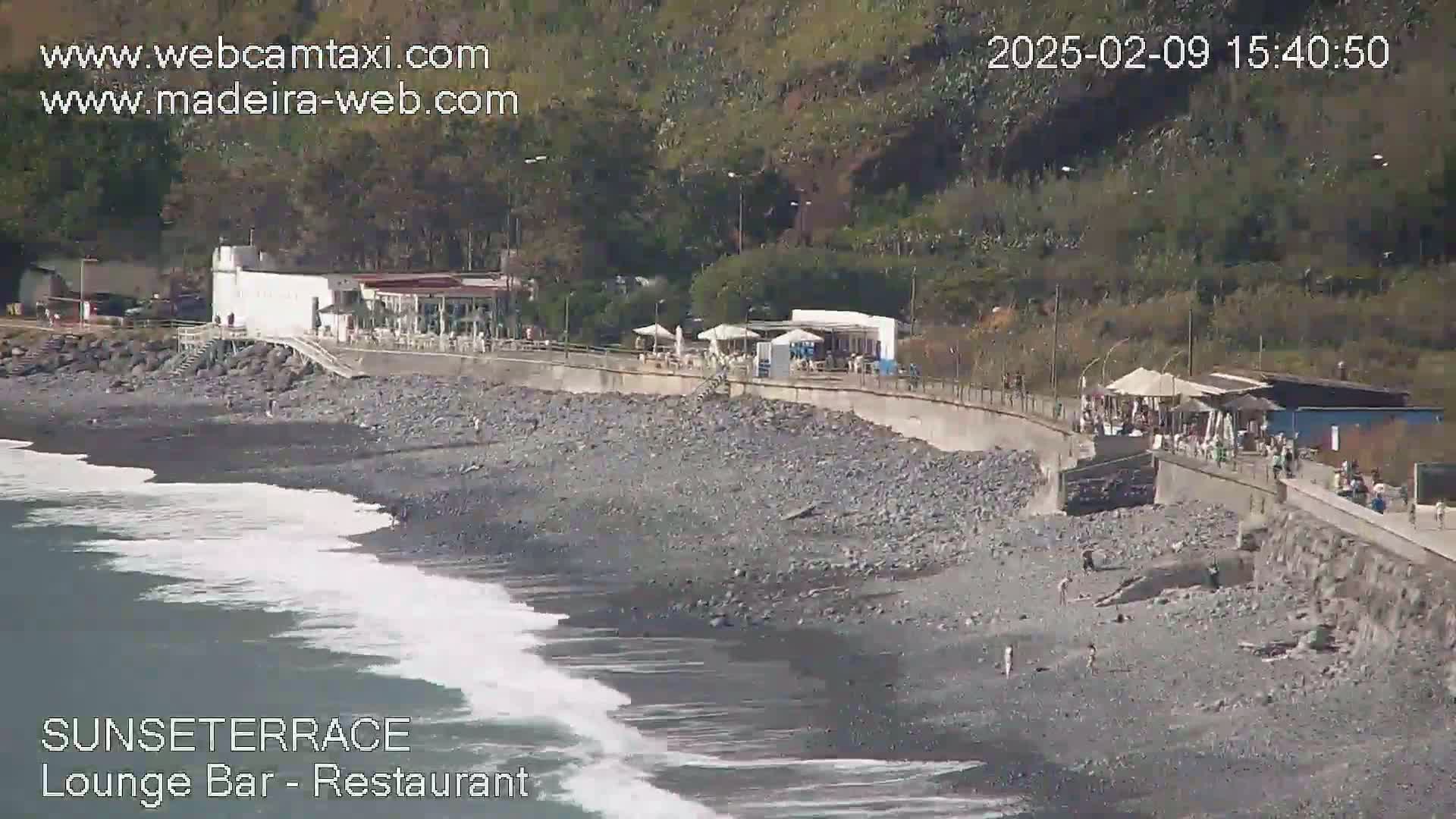 A rocky beach with a restaurant and a seawall is shown, with gentle waves lapping the shore on a sunny day.