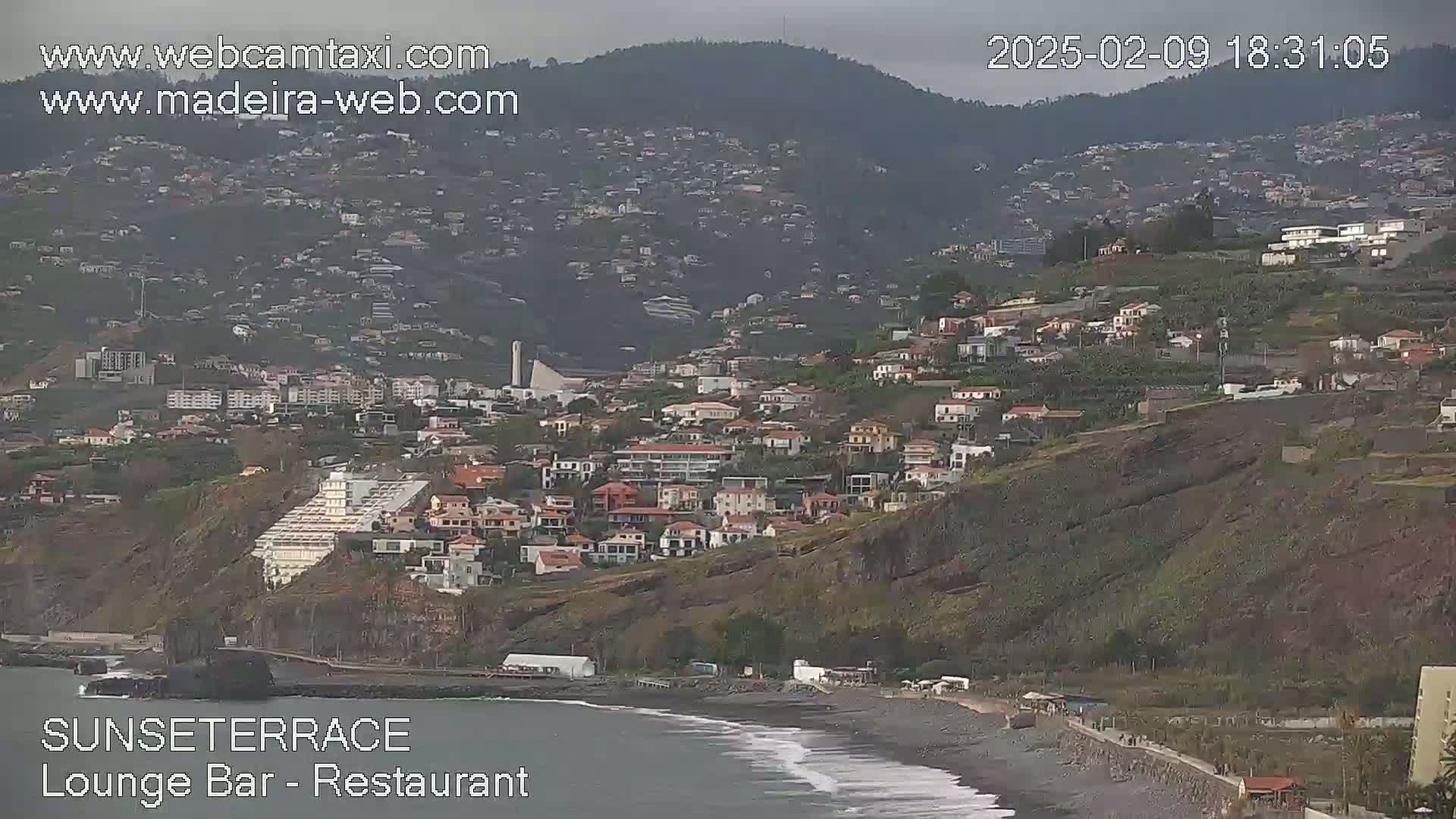 A coastal town nestled on a hillside overlooking a dark-sand beach under an overcast sky.