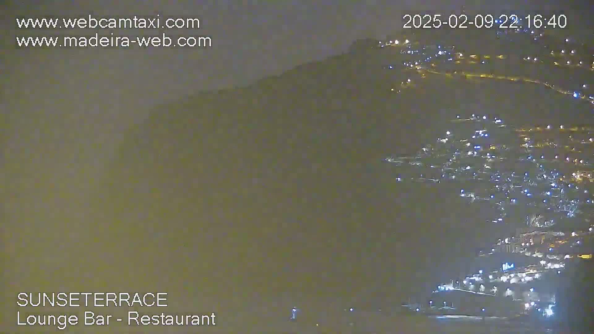 A hazy night scene shows a hillside town with numerous lights illuminated against a dark, overcast sky.